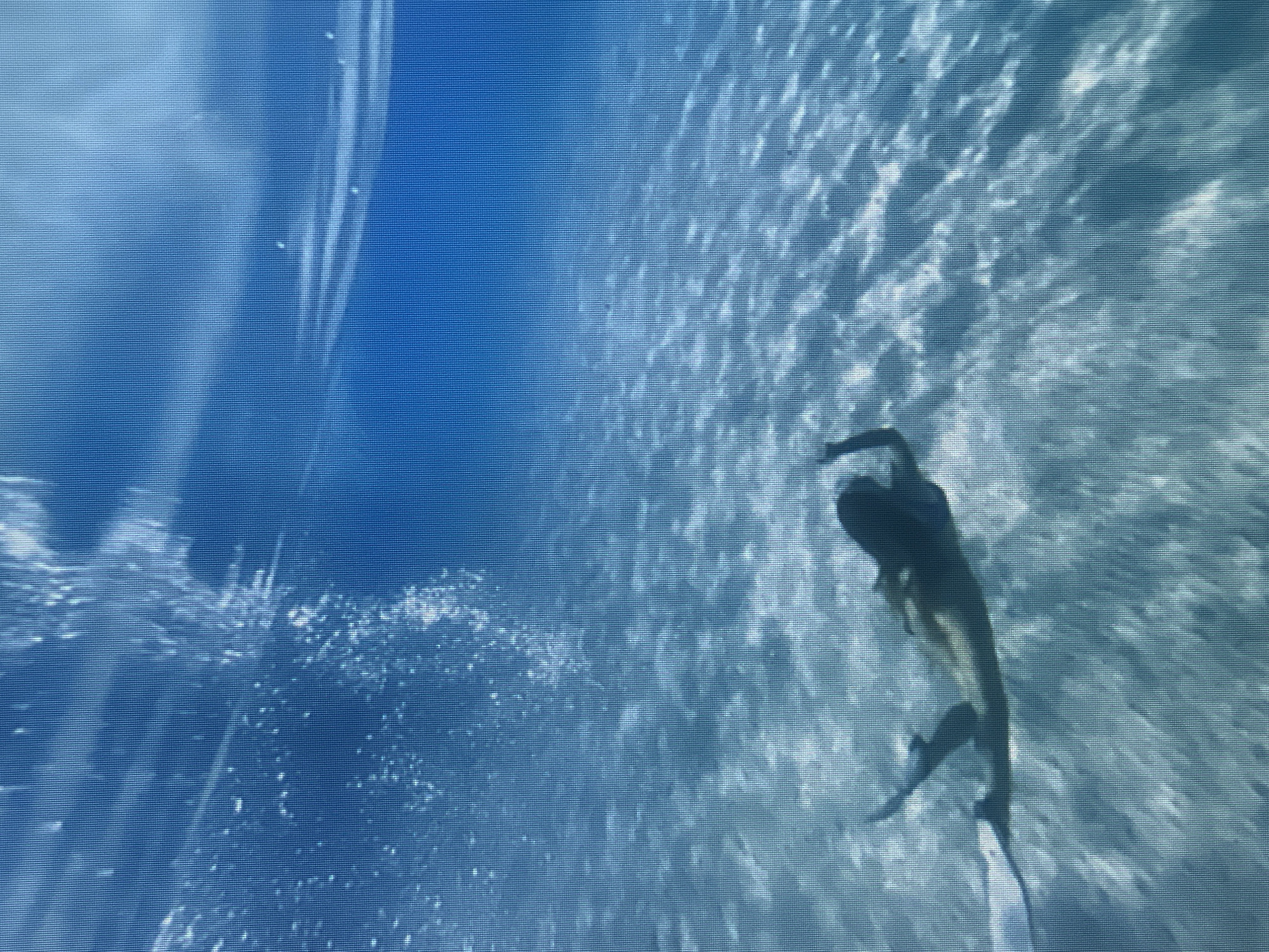 An underwater shot shows a person swimming in clear blue water over a sandy bottom. The person is in a horizontal position, with their arms extended and legs kicking, propelling them forward. Sunlight filters down from the surface, creating dappled patterns on the sand. The water appears shallow and calm. The location is Unknown, Unknown.