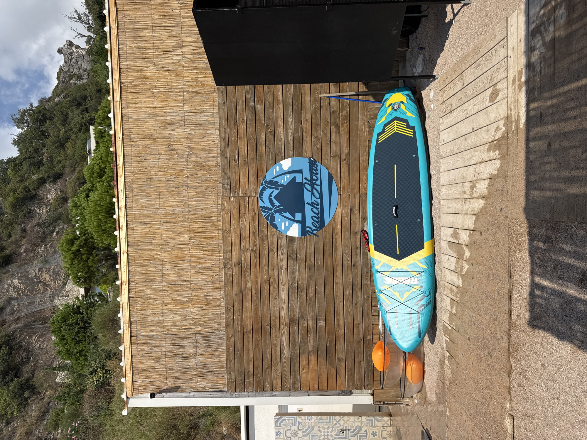 The image shows a stand-up paddleboard resting on a wooden deck in front of a building with a bamboo facade and a wooden wall. A circular blue sign with the words "Beach House" and an illustration of palm trees and a hut is mounted on the wooden wall. Two orange buoys are visible behind the paddleboard, resting in clear containers. To the right, a large black speaker stands next to the wooden wall. The background features a rocky hillside covered in dense green vegetation and a partly cloudy sky. The ground is a mix of sand and gravel, with wooden planks forming a path. The scene appears to be located in Mirište, Montenegro.
