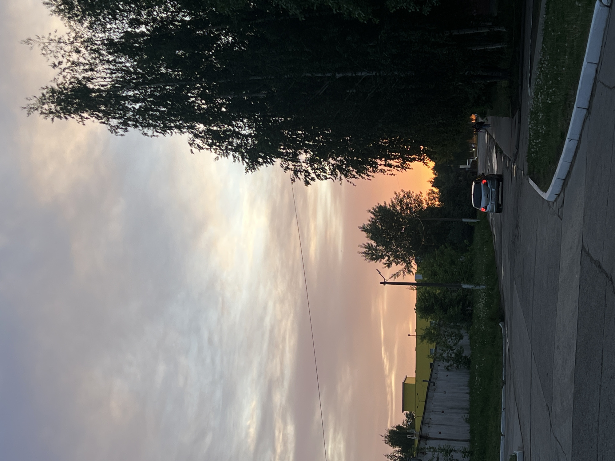 The image shows a street scene in Chaikovsky, Russia, likely taken during a sunset. A grey car is parked on the right side of the road. A person is walking on the sidewalk in the distance, partially obscured by trees. The sky displays a gradient of colors ranging from light grey to orange, indicative of the sunset. Overhanging tree branches frame the top of the image, and buildings are visible in the background on the right. A power line stretches across the scene.