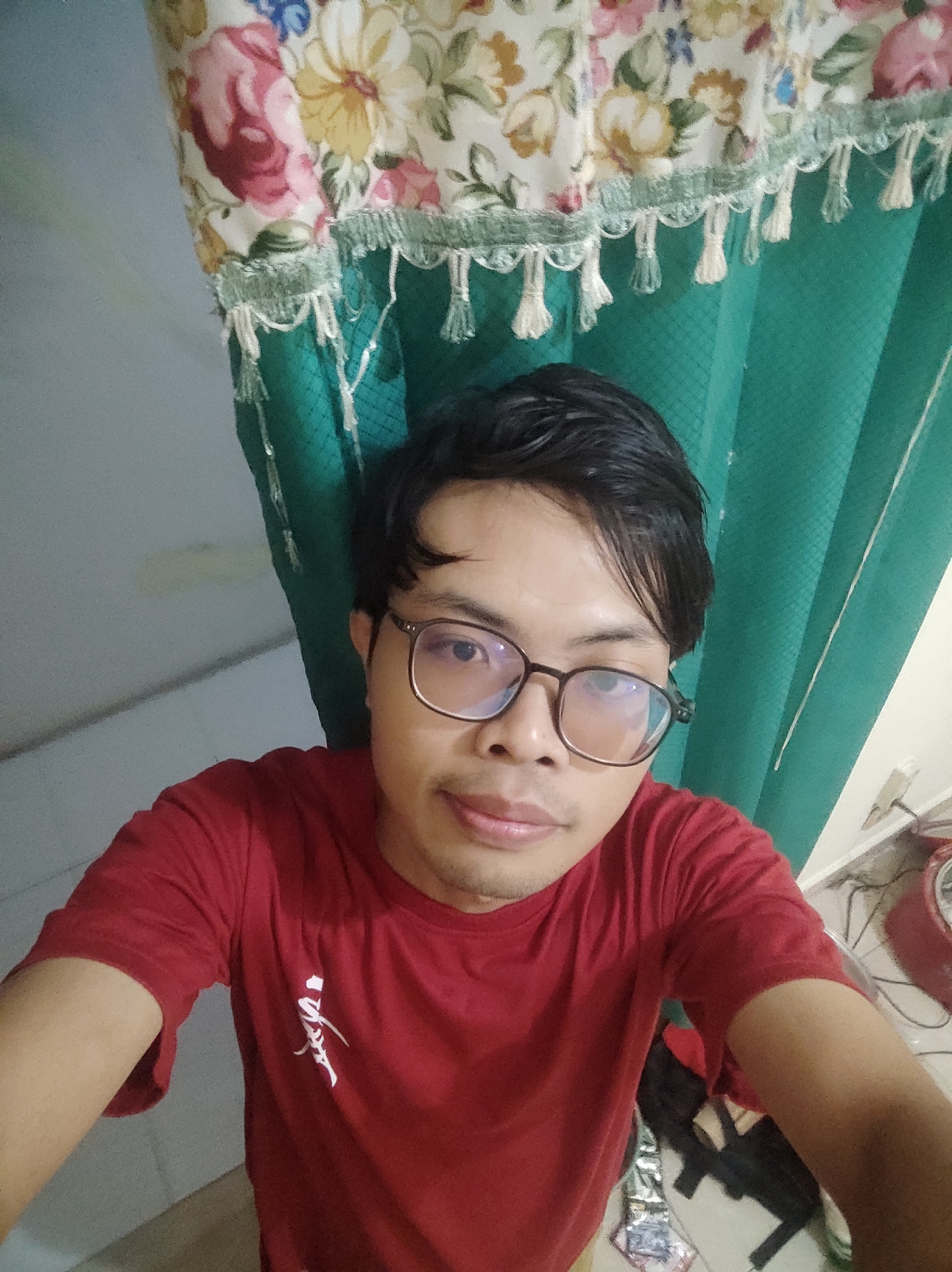 A young man with dark hair and glasses is taking a selfie in what appears to be a room. He is wearing a red t-shirt with a white graphic design on the left side of his chest. Behind him, a green curtain is visible, and above that, a valance with a floral pattern and fringe. The background also shows a light-colored wall and a floor. There are electrical cords and a red circular object on the floor to the right. The image is a close-up of the man's upper body. The location is specified as Trihanggo, Indonesia.