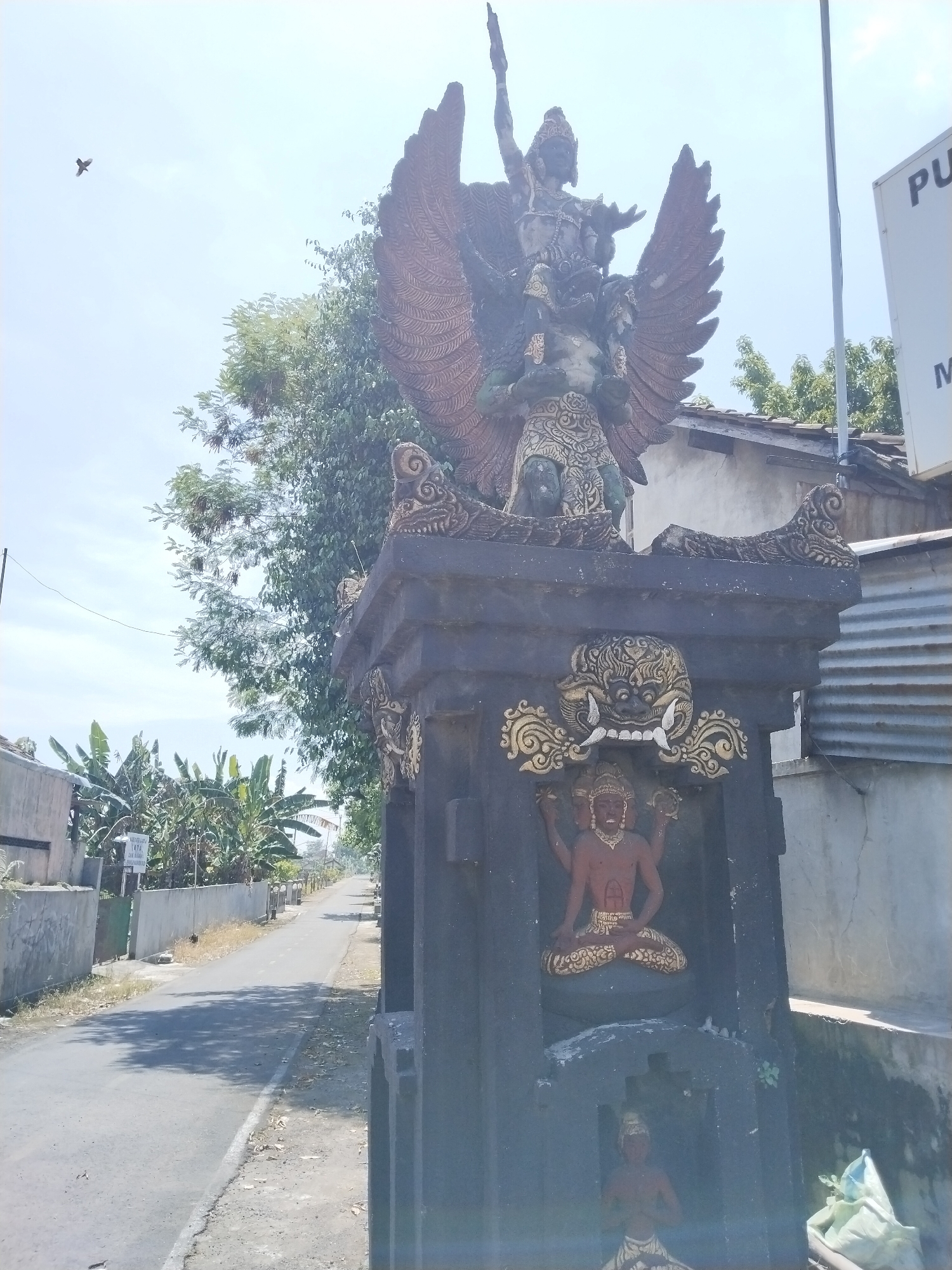 The image shows a roadside monument in Masaran, Indonesia. The monument is a dark gray structure with gold accents. It features three distinct sculptural elements:
1. At the top is a depiction of Garuda carrying Vishnu, a common motif in Indonesian art and representing power, speed, and strength.
2. Below that is a carving of a Kala, a mythical creature associated with time and destruction, often found above doorways to ward off evil.
3. On the main body of the monument are depictions of a deity with four heads.
The monument sits on a road in a suburban or rural area. There is vegetation along the roadside, and simple dwellings are visible in the background. The sky is bright, suggesting a sunny day. A bird is visible in the sky to the left.