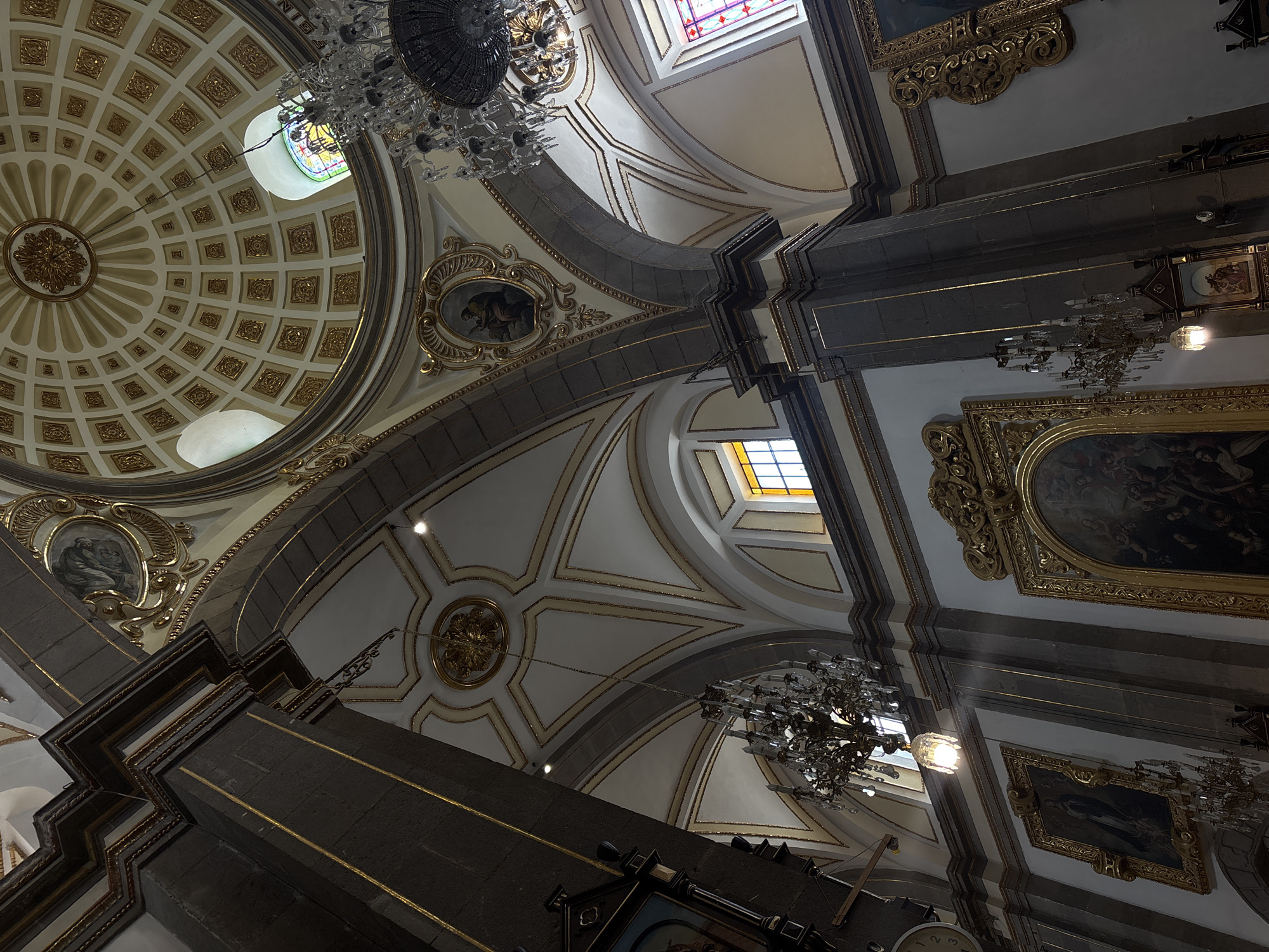 The image captures an interior view of a richly decorated church, likely located in Puebla, Mexico, judging by the architectural style. The camera is pointed upwards, emphasizing the intricate ceiling and dome. The ceiling features a complex design of interlocking geometric shapes, adorned with gold accents. A large dome is present on the left side of the frame, decorated with a grid pattern and a central rosette. Two ornate chandeliers hang from the ceiling, providing illumination. There are also framed paintings and decorative elements along the walls. The architecture displays a mix of Baroque and Neoclassical styles, typical of Mexican colonial churches. The overall impression is one of grandeur and opulence.