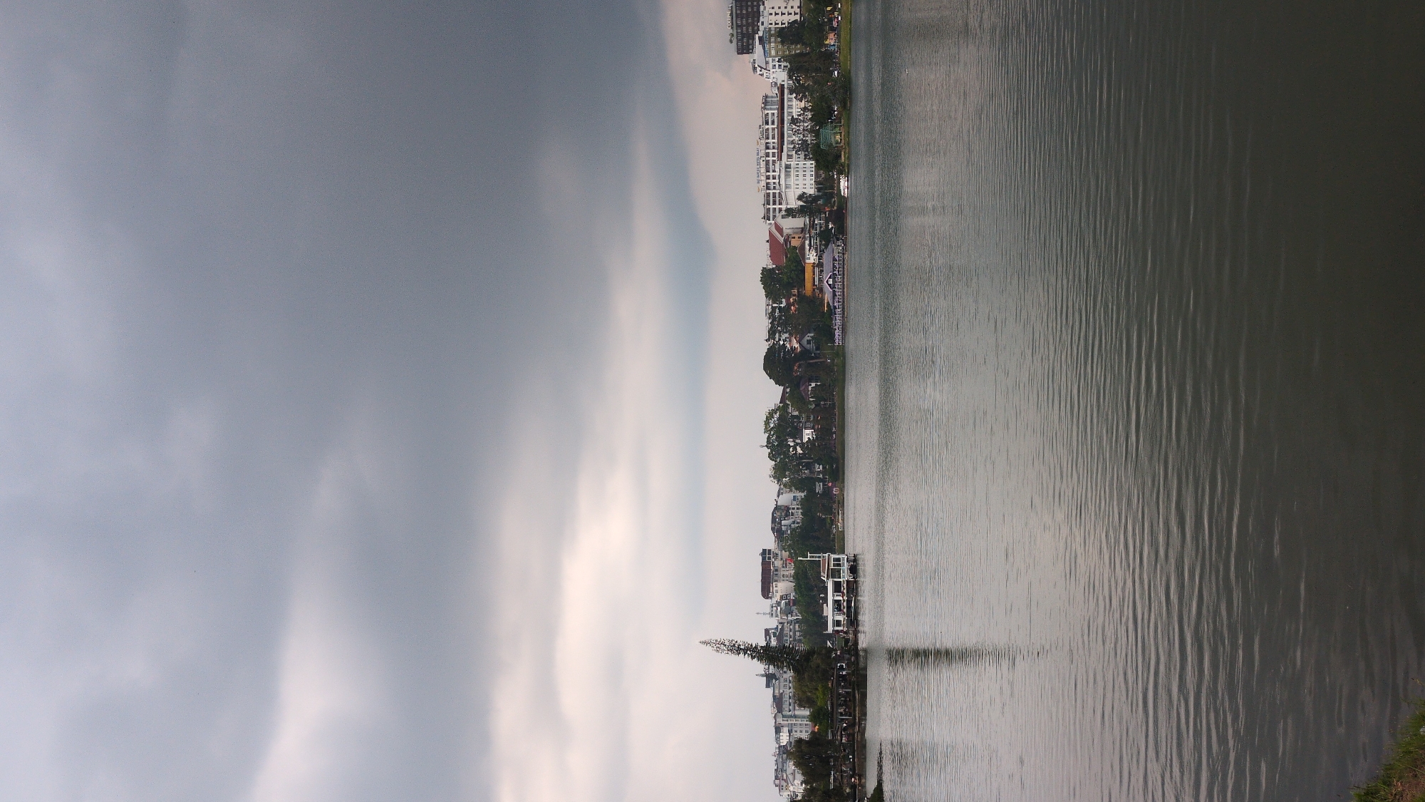 The image shows the city of Da Lat, Vietnam, as seen from across Xuan Huong Lake. The lake occupies the right side of the image, with a slightly rippled surface reflecting the sky and the city skyline. The city stretches along the shore, featuring a mix of low-rise buildings and taller structures. The sky on the left is overcast with heavy, grey clouds, indicating possible rain. The perspective is from the water level, looking towards the city. There are no people visible in the frame.