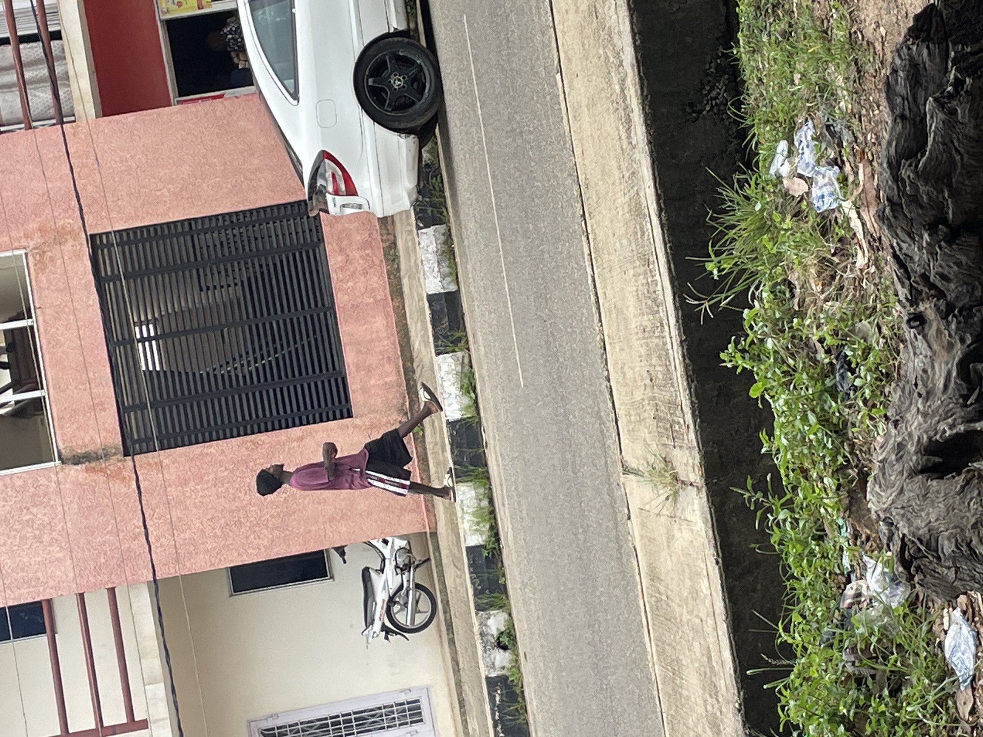 In Jos, Nigeria, a young man wearing a purple t-shirt, black shorts with white stripes, and a black beanie walks on a sidewalk next to a pink building. He is walking towards the right side of the frame. To his left, a white motorcycle is parked on the sidewalk. A white car is parked on the street to his right. The sidewalk is bordered by a curb with black and white markings. The street is paved and has white lines indicating lanes. The foreground of the image shows a patch of overgrown grass with some litter and a large piece of wood. The background features the pink building with windows and balconies.