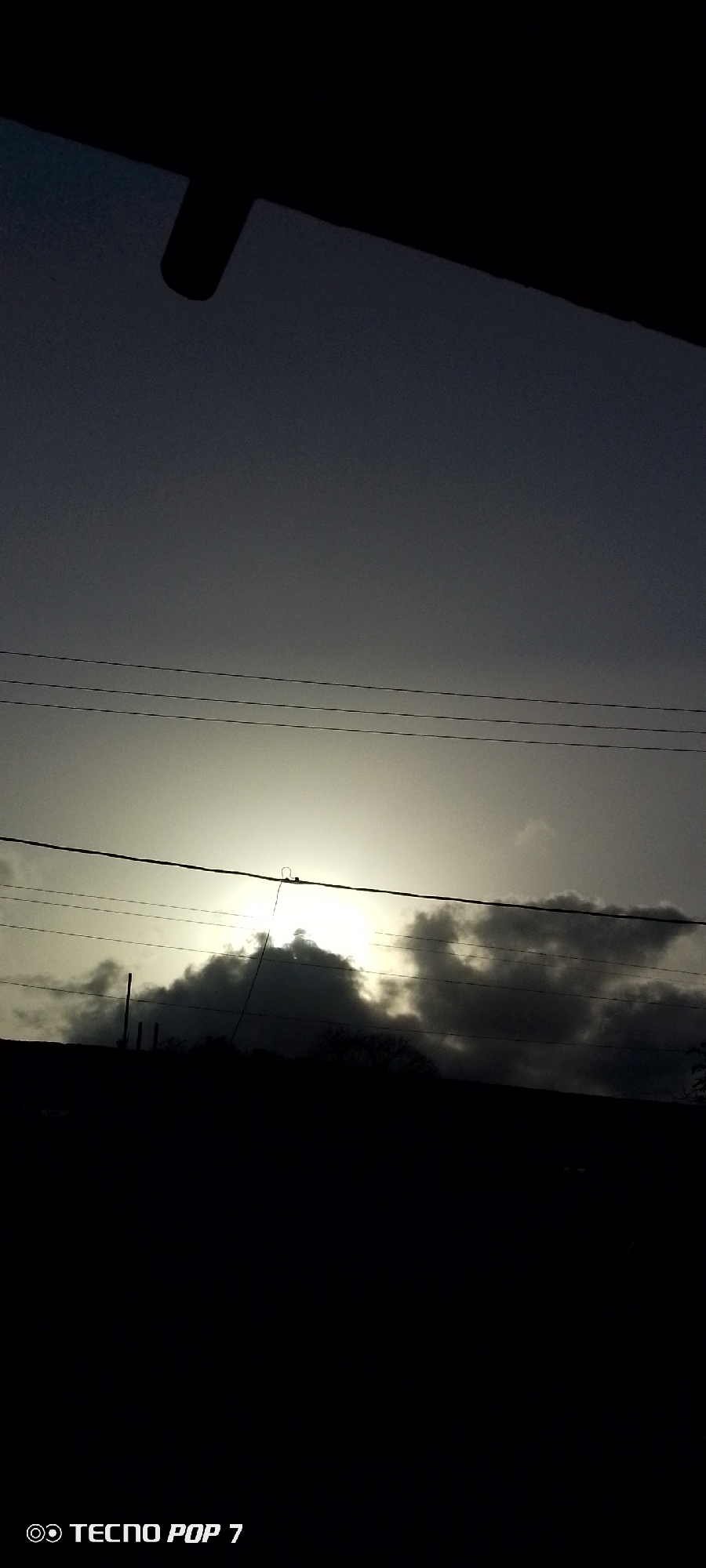 A low-angle shot of a cloudy sky, possibly at dusk or dawn. Silhouetted buildings or trees are in the foreground, obscuring the lower portion of the image. Power lines cut across the sky horizontally. The sky is mostly gray, with a bright, indistinct area possibly indicating the sun behind the clouds. The bottom of the photo contains the text "TECNO POP 7", indicating the phone model used to take the photo. The location is assumed to be in the city of Altagracia, Venezuela.