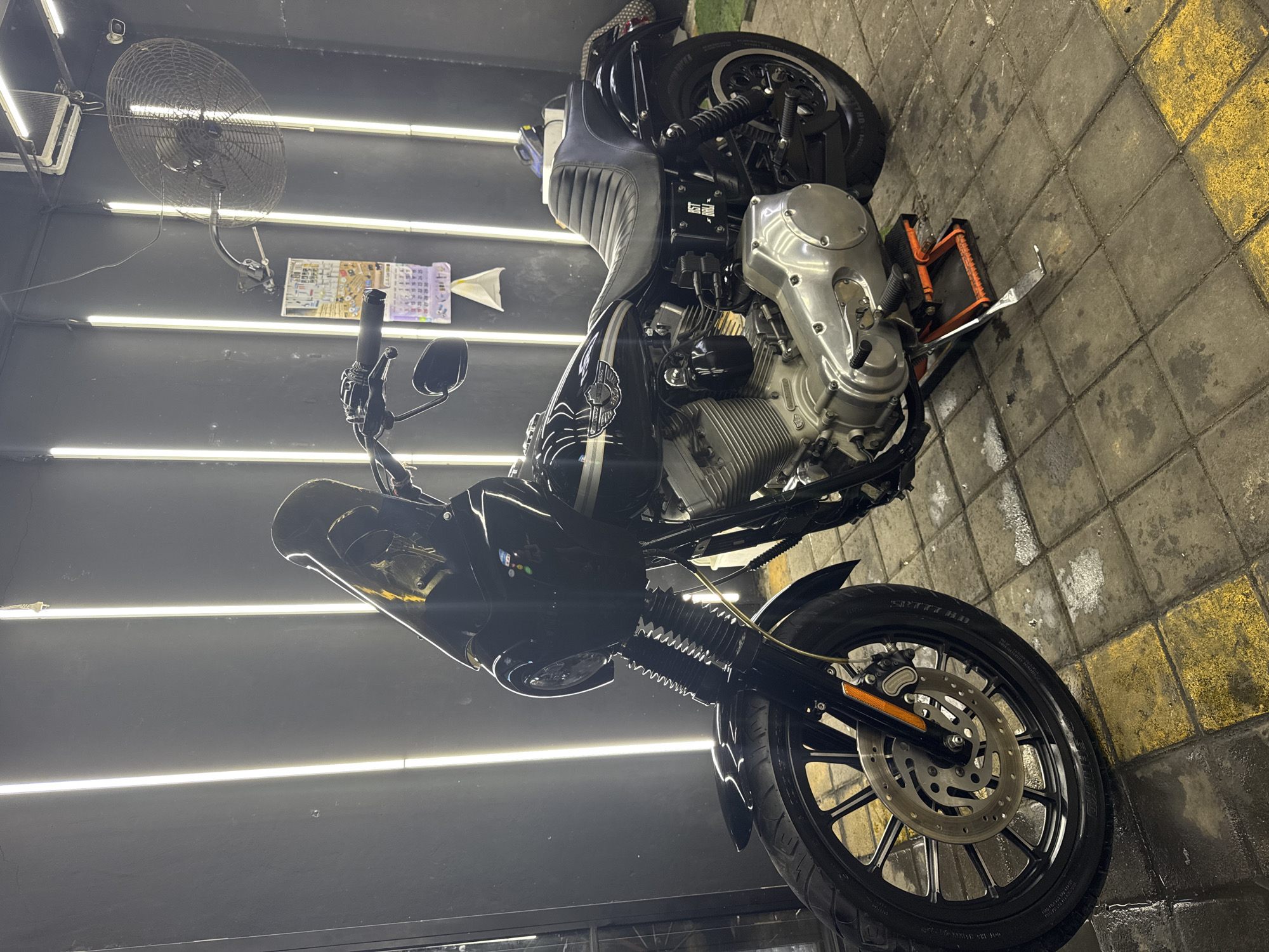 A slightly rotated close-up shot shows a motorcycle inside a repair shop in Ungasan, Indonesia. The motorcycle, primarily black and silver, rests on an orange stand atop a tiled floor with alternating gray and yellow blocks. The front wheel faces the viewer, showcasing the tire brand “SPIRIT HD.” The back wheel is also visible, though more obscured. The motorcycle's engine is a prominent silver hue. Overhead, a black wall-mounted fan is positioned near a calendar hanging on a black wall. Above, horizontal strip lighting illuminates the scene. The setting suggests an automotive or motorcycle service facility.