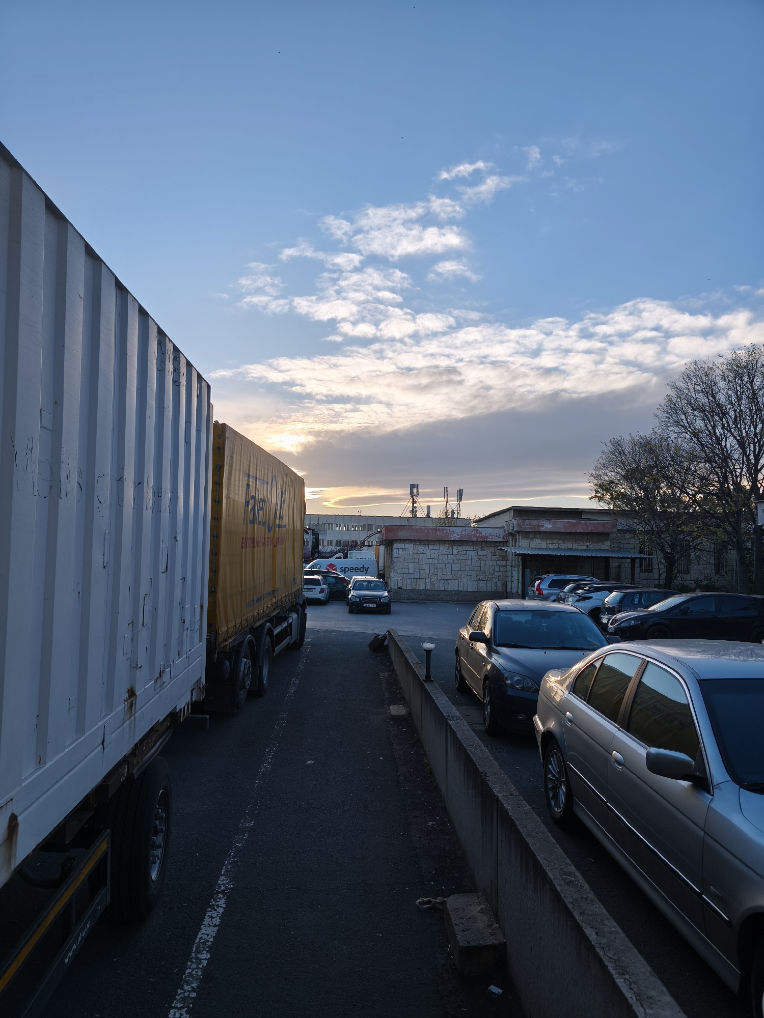 This media file depicts an outdoor scene, likely a parking area or industrial yard, at either sunrise or sunset. The sky displays a gradient of colors, from clear blue at the top to warm orange and yellow near the horizon, with scattered white and grey clouds.
In the left foreground, a large white cargo truck or trailer dominates the frame, its ribbed side showing signs of wear and rust. Faint, weathered text is visible on its side, and the URL "www.d-i.eu" can be seen on its chassis. Immediately to the right, a yellow curtain-side truck is parked, bearing the "PALETOL" logo and Cyrillic text, including "ЕКСПРЕСНА ЛИНИЯ ДО" (Express Line to).
The mid-ground features several parked cars, including a silver sedan, a dark grey sedan, and other vehicles. A white van with the "Speedy" logistics company logo is also visible. Some car license plates, such as one starting with "CB" (Sofia, Bulgaria registration code), provide location context for Bulgaria.
In the background, utilitarian buildings with stone or concrete facades are visible, and several antenna towers stand on the roof of one structure. Bare-branched trees are silhouetted against the sky on the right. A low concrete barrier runs along the right side of the road, separating it from an elevated parking area. The scene is devoid of people, conveying a quiet, transitional time of day in a commercial or industrial setting, consistent with Burgas, Bulgaria.