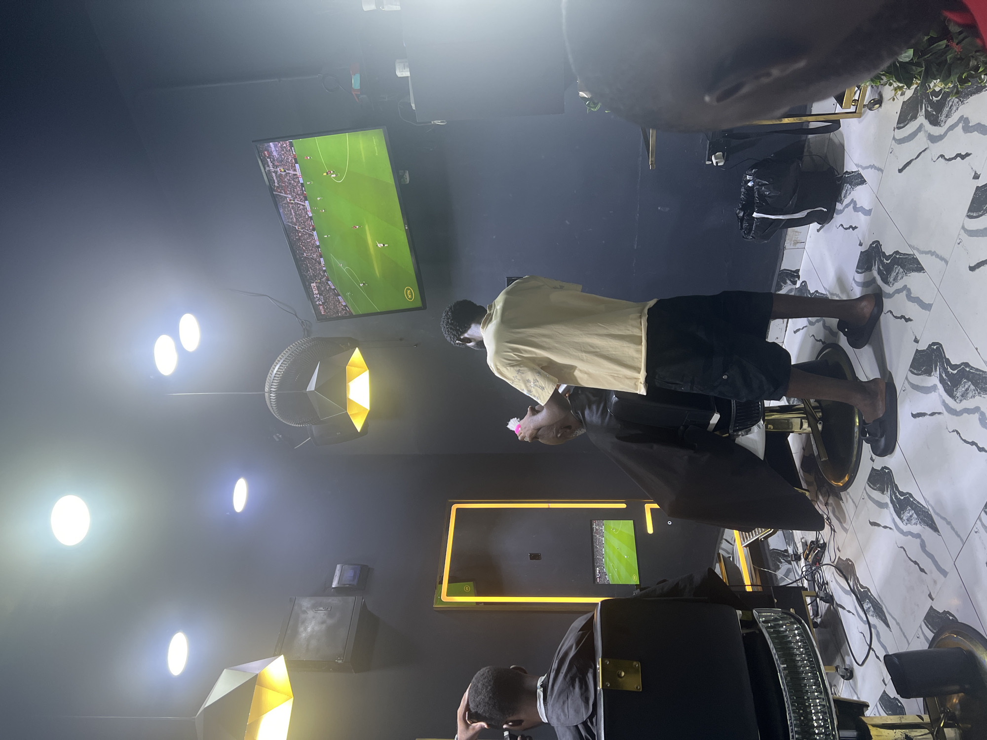 The image depicts the interior of a barber shop, likely located in Lugbe, Nigeria, evidenced by the general setting and ambiance. The scene is oriented in a portrait layout, and the angle is slightly tilted. A barber, dressed in a yellowish shirt and dark shorts, stands beside a client who is seated in a black barber chair with a cape draped over them. The barber is in the midst of giving a haircut.
There are at least two televisions visible. One is playing a soccer match. The shop has several lights, including uniquely shaped decorative lights. The walls are painted dark, and the floor has a white and gray patterned design. A second client waits in another barber chair. The environment shows a modern yet simple shop design. Additional barber equipment, including tools and a fan, are present.