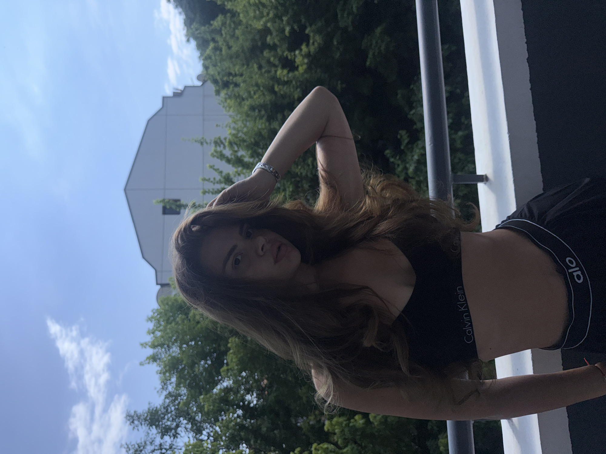A young woman with long, brown, wavy hair is standing on a balcony with a railing in the foreground. She is wearing a black Calvin Klein bra and black shorts with "alo" written on the waistband. She has a bracelet on her right wrist. Behind her are green trees and a white building. The sky is blue with some white clouds. The scene appears to be in a residential area of Plovdiv, Bulgaria.