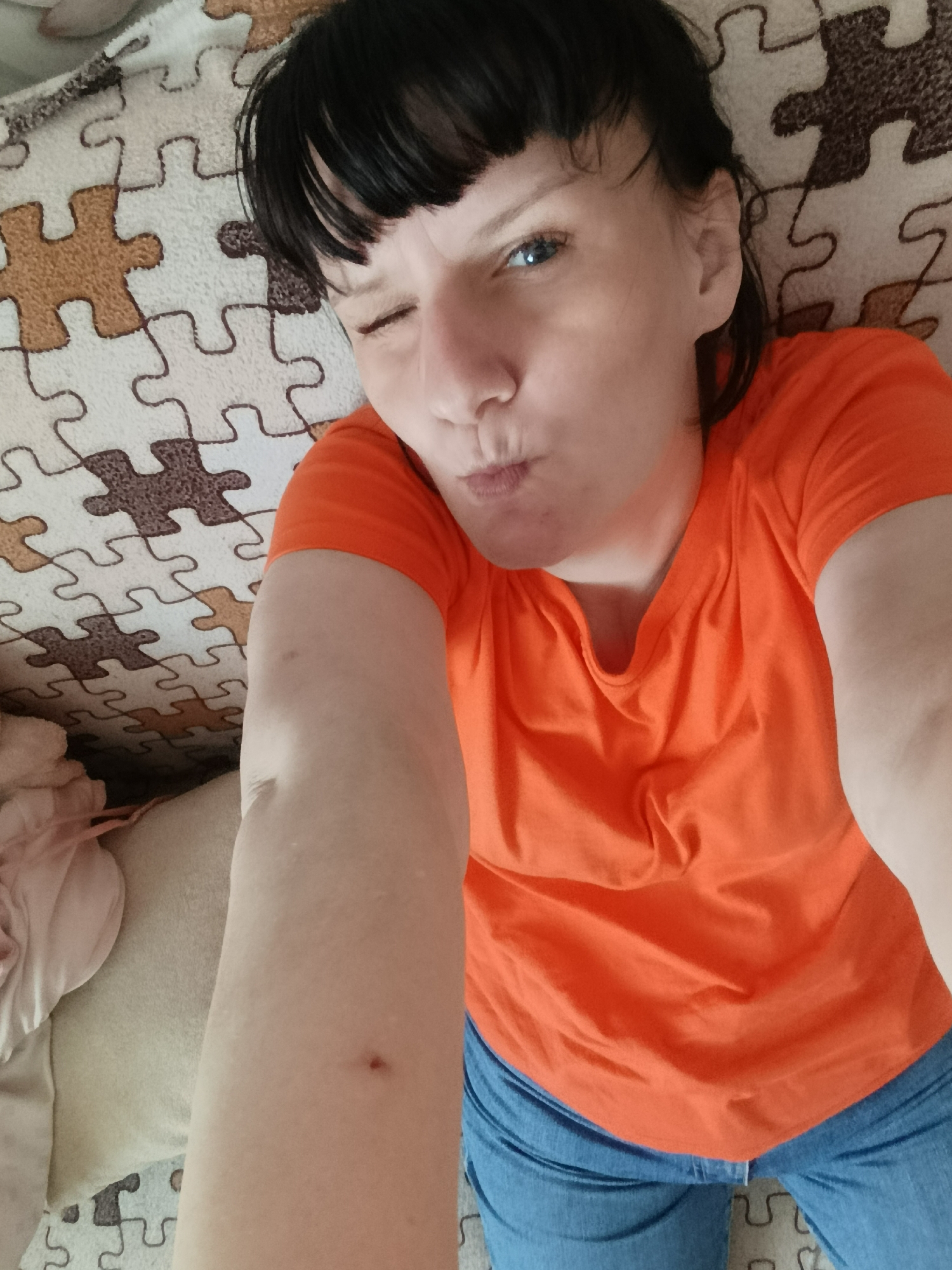 A woman is lying on a puzzle mat while winking at the camera. She has short, dark hair and bangs, and is wearing an orange T-shirt and blue jeans. The background includes the puzzle mat, a pillow, and a blanket. There is no information to indicate the location is Фоки, Russia.