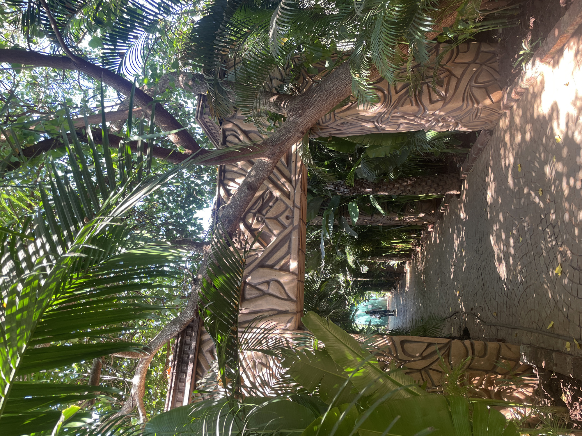 A sunny day in Chopdem, India, reveals a view through an ornate, patterned stone archway overgrown with dense tropical vegetation. Palm fronds are prominent in the foreground, framing the scene. The archway leads onto a paved pathway that extends into the distance, partially shaded by foliage. Two figures are faintly visible in the distance along the pathway. The architecture of the archway features intricate carvings in a light, earthy tone. Trees appear to be growing through and around the archway, further integrating the structure into the surrounding natural environment.
