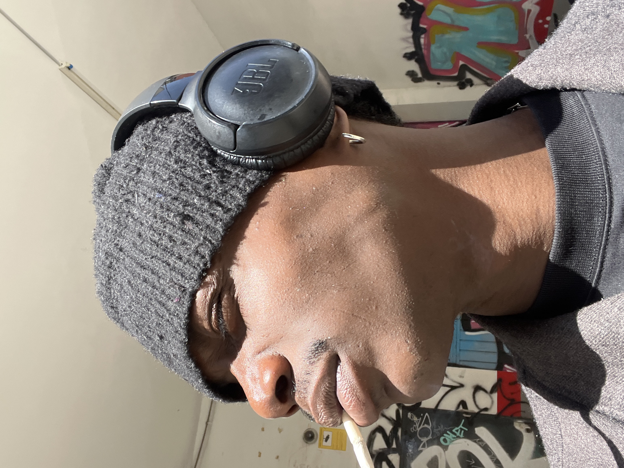 A close-up shot, taken from a low angle, shows a person's face in profile. The person has dark skin, a knit cap, and black JBL headphones. A cigarette is held in the person's mouth. A small hoop earring is visible. The background shows graffiti-covered walls, suggesting an urban setting. The location is Barcelona, Spain, based on the graffiti style and architectural details.
