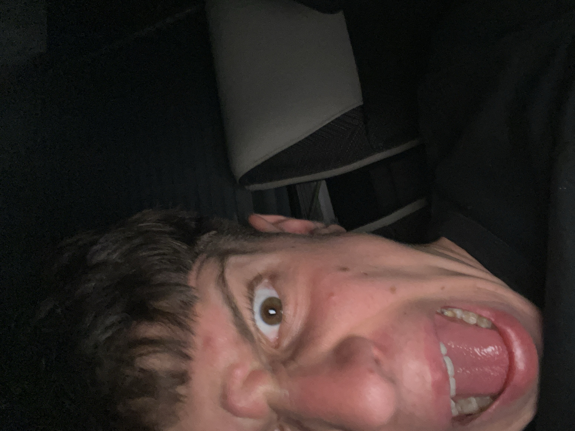 A selfie shows a person's face at an extreme upward angle, mouth wide open, revealing teeth and tongue. The person has brown hair and one visible brown eye. The background is dark, with part of a gray and black chair visible. The location of the image is unknown and is not Saraia, Bulgaria.
