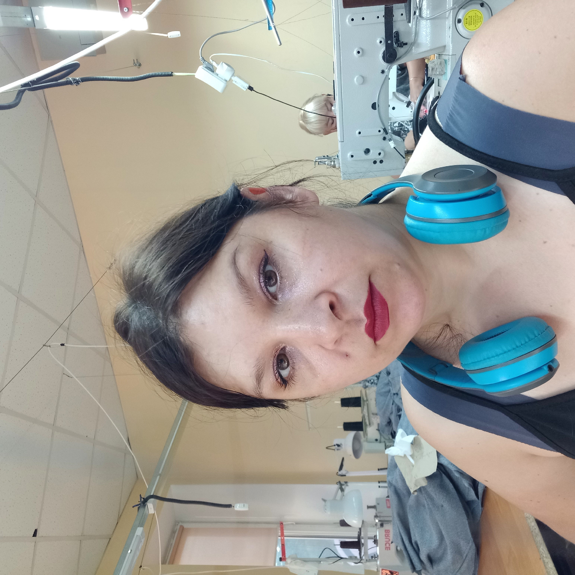 The image shows a woman with dark hair and headphones around her neck, looking at the camera. She is wearing makeup and has dark red lipstick on. She is in what appears to be a sewing workshop, with sewing machines and fabric visible in the background. Another woman with blonde hair is visible in the background, working at a sewing machine. The environment suggests an industrial or commercial setting, possibly a factory or a small business. The overall lighting is bright and artificial, consistent with an indoor workspace. The image is a selfie, taken by the woman in the foreground. It is not possible to determine the exact location within Kropyvnytskyi, Ukraine from the provided image.