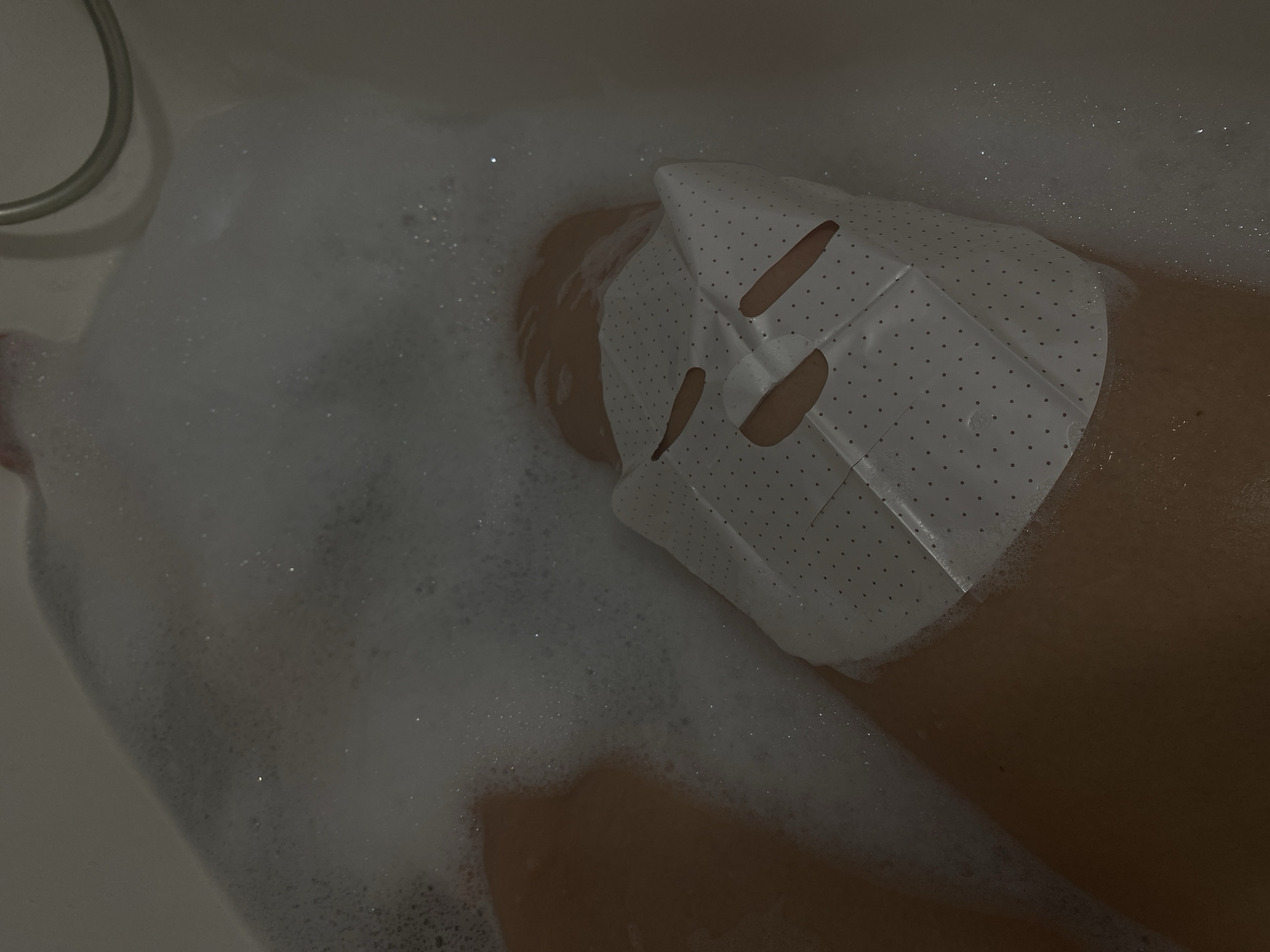 The scene depicts an individual undergoing a personal care routine within a bathtub, located in the city of Sochi, Russia. The perspective is first-person or close overhead, looking down into the tub. The environment is dimly lit, suggesting an indoor bathroom setting. A person's upper torso and face are visible, submerged in a bathtub filled with water and a significant amount of white foam bubbles. A white sheet mask, speckled with small dark dots and featuring cut-outs for eyes, nose, and mouth, is applied to the individual's face. The mask is slightly folded at the chin and nose areas. A chrome or silver-colored flexible shower hose is visible in the upper right quadrant, draped over the edge of the tub. The bathtub itself appears to be a light, possibly off-white, ceramic or porcelain material. No other individuals are present or discernable. The primary action is the individual bathing and applying a cosmetic face mask.