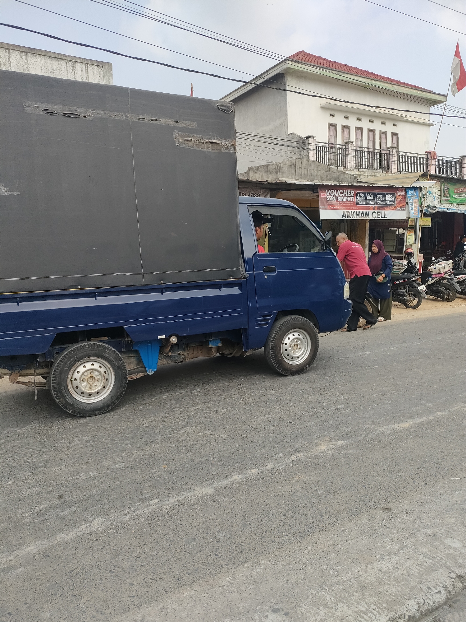 In Sukoyoso, Indonesia, a blue pickup truck with a black tarp covering its cargo bed is parked on an asphalt road. The truck is positioned at an angle, facing slightly to the right of the frame. Two individuals are interacting near the passenger side of the truck. A man in a red shirt and black pants is leaning towards the truck, appearing to speak with or assist someone inside. Beside him, a woman in a dark hijab and green skirt stands with her hands clasped, looking towards the truck. Several motorcycles are parked along the sidewalk to the right of the truck. In the background, a two-story white building with a red tiled roof stands. An Indonesian flag is visible flying from the building. Signs for "ARKHAN CELL" and mobile data package promotions are displayed on the storefronts. The sky is overcast.