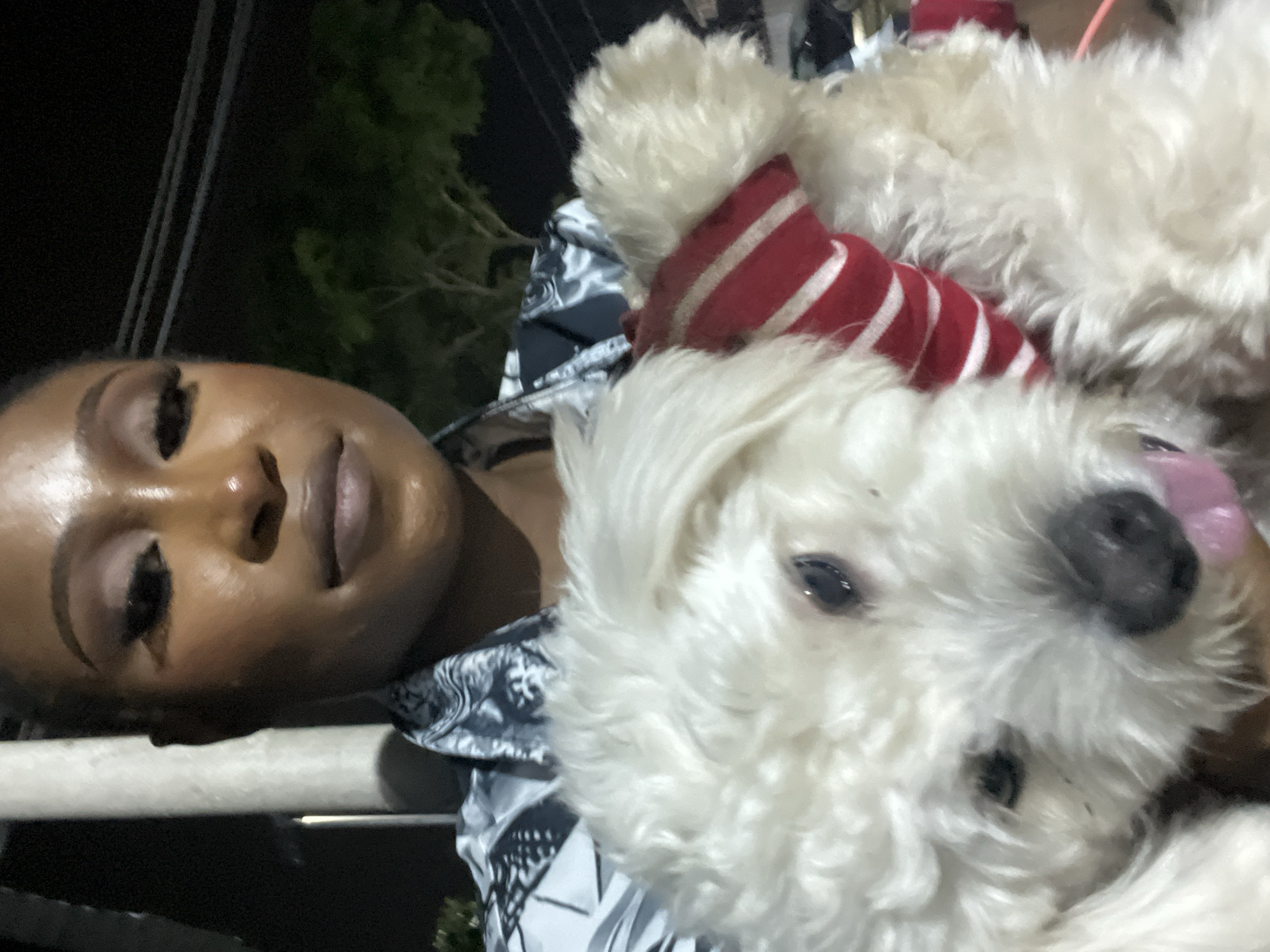 A close-up, waist-up, eye-level shot features a Black woman holding a white fluffy dog outdoors at night. The woman, facing the camera, wears a black and white patterned shirt and eye makeup. The dog, positioned in front of the woman, is wearing a red and white striped scarf. The background is dark, with hints of trees and possibly a pole. The overall impression is a casual, outdoor setting at night.