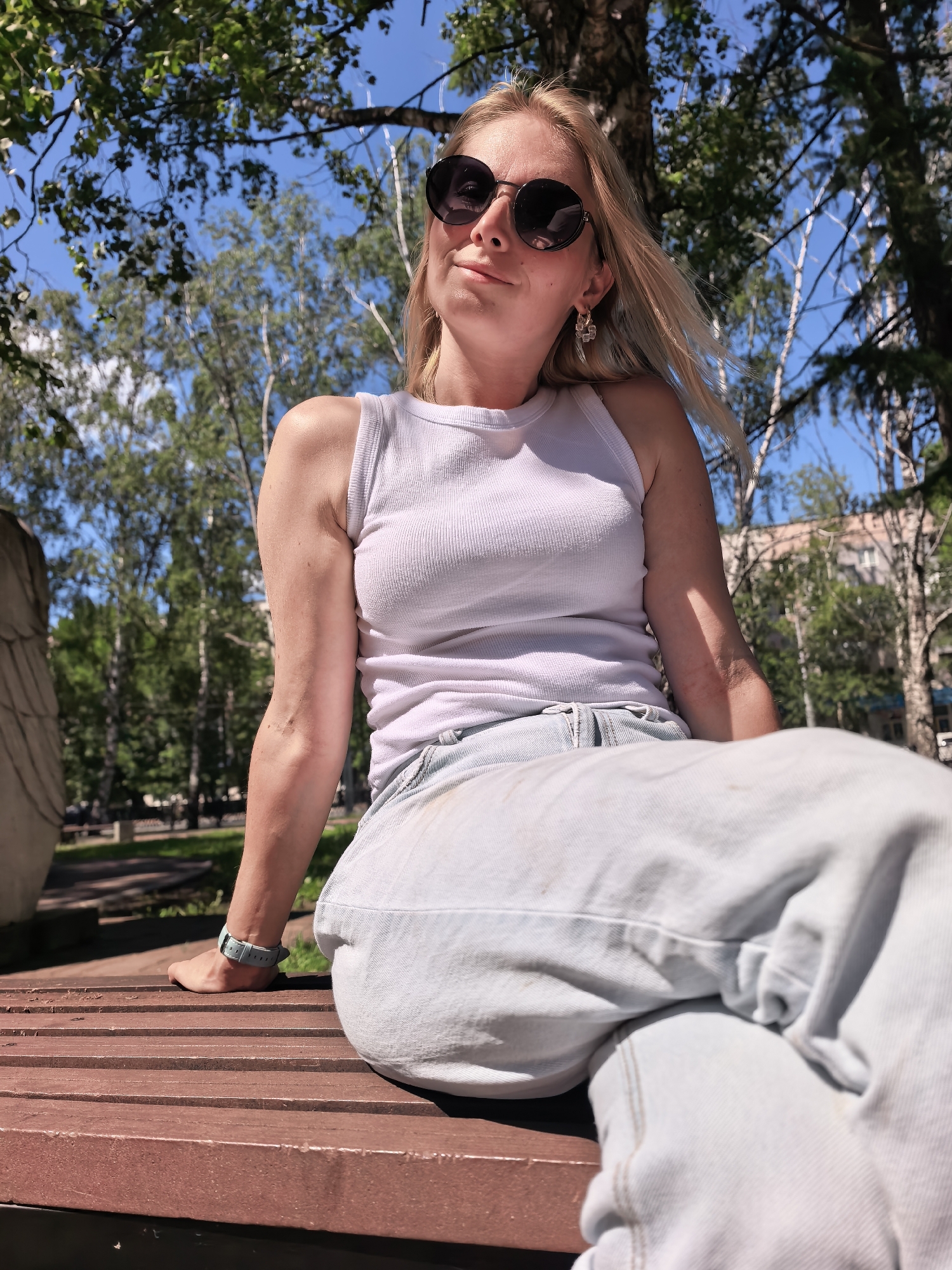 A young woman with blonde hair, wearing a white tank top, light blue jeans, and black sunglasses, sits on a wooden bench outdoors. She is looking up and to her left, with a slight smile. The background features trees and a building, suggesting an urban park setting. The lighting indicates a sunny day. There is no specific visual information in the image to confirm the location as Sterlitamak, Russia.