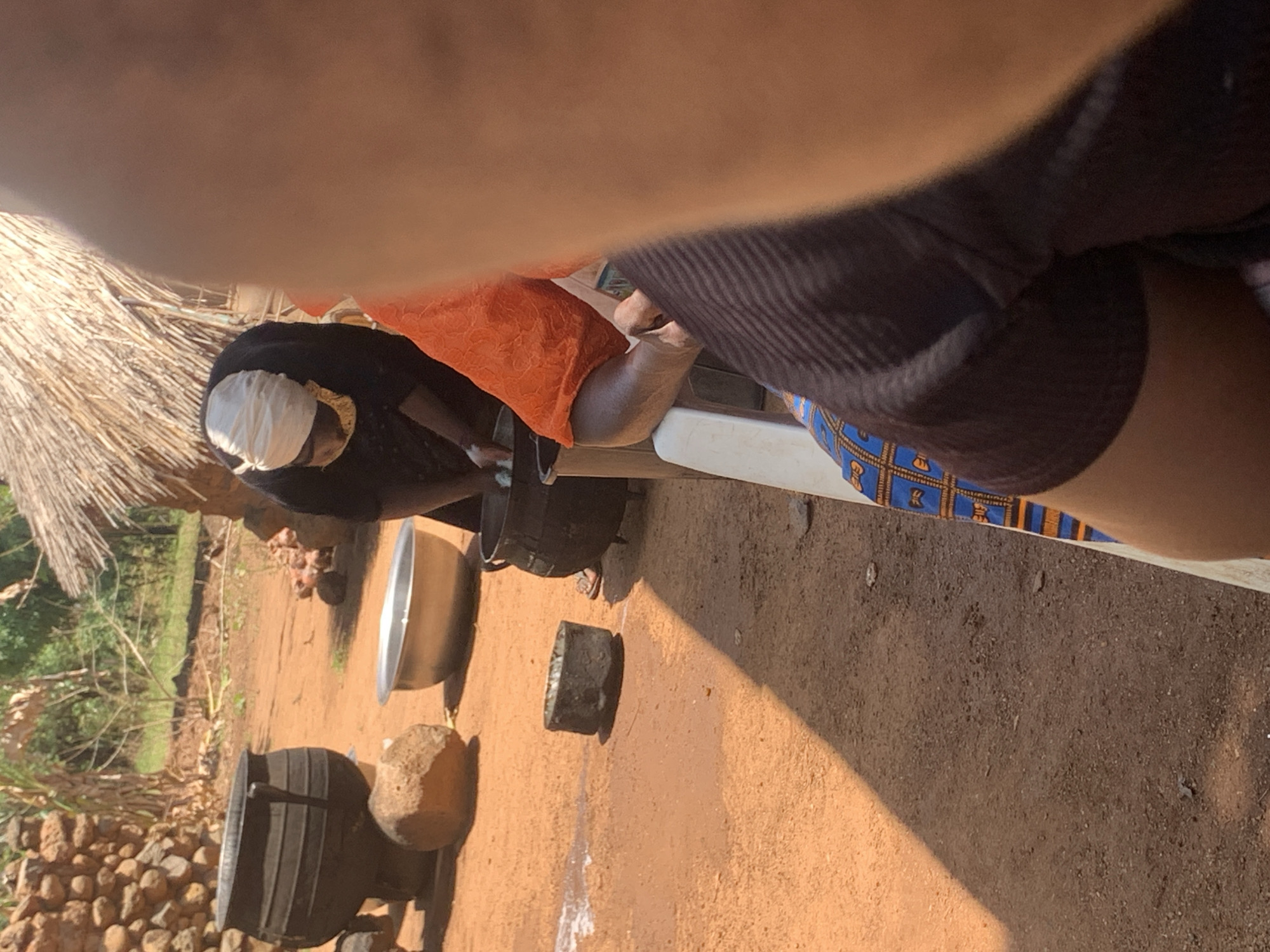 In Fada, Nigeria, the image shows an outdoor scene with a person wearing a black shirt and patterned blue and orange shorts sitting on a white chair. An individual wearing an orange shirt and a white head covering is bent over a large black pot. Next to the pot is a metal bowl, a piece of wood, and a large rock. The ground is brown dirt, and there is a thatched roof in the background. A stone wall and trees are visible in the distance.