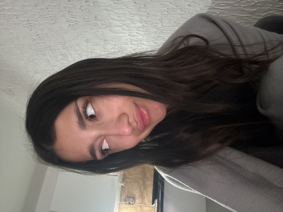 A young adult female is depicted in a close-up indoor shot, understood to be located in Zankovići, Montenegro. The individual has long, dark hair, parted centrally, cascading over her shoulders. Her face is turned slightly to the left, with her eyes wide open and directed upwards and to the viewer's right. Her lips are lightly pursed. She is wearing a dark garment and is partially enveloped in a light grey blanket or shawl draped over her left shoulder and chest. The background on the left side features light brown/tan wall tiles above a dark countertop or surface. The right background consists of a textured, off-white wall, possibly stucco or plaster. No other individuals or direct interactions are visible.