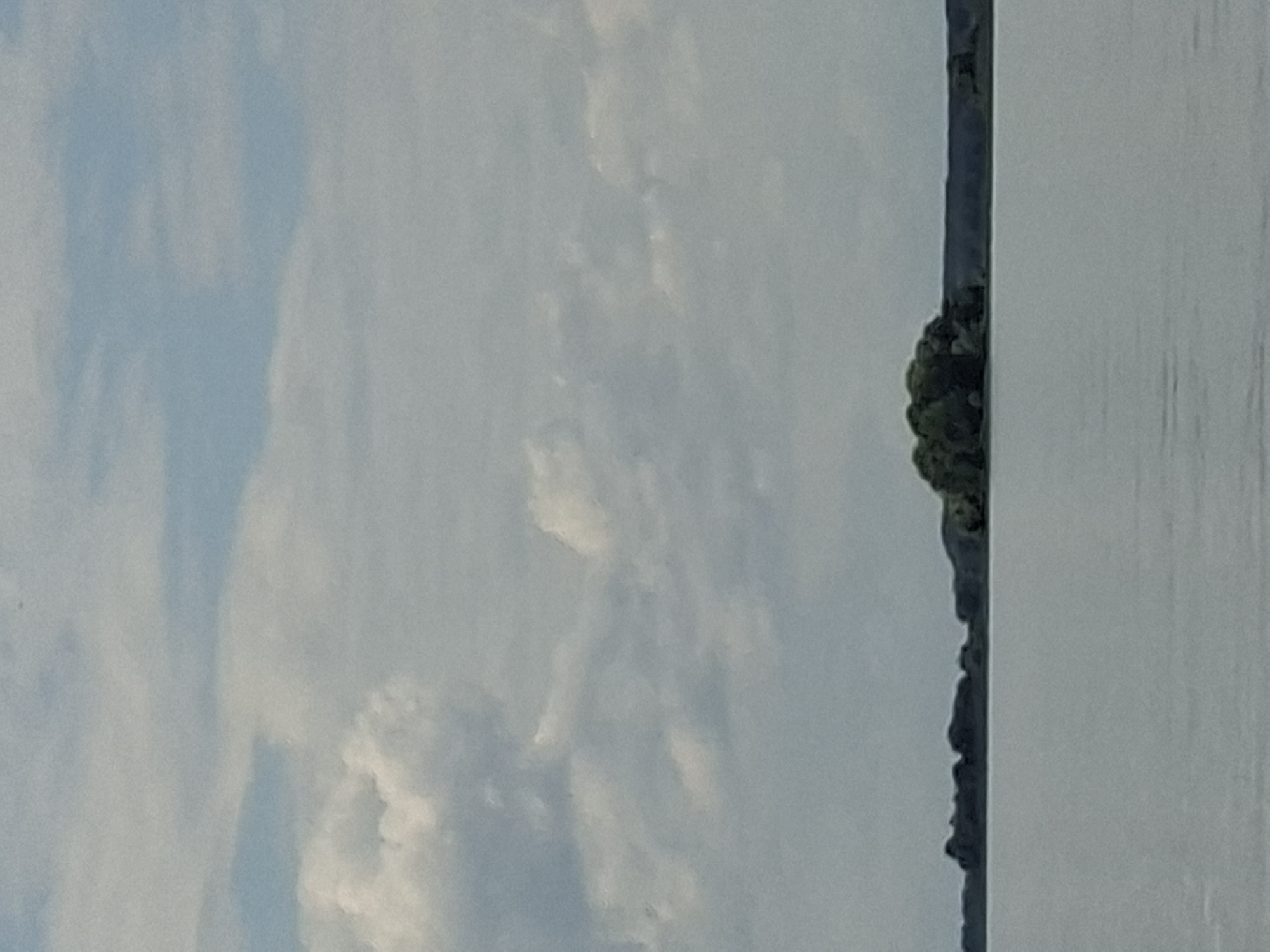 A landscape scene depicts a body of water with a tree-covered island in the distance, set against a sky filled with clouds. The water is a muted gray, reflecting the overcast sky. The island appears small and dense with foliage, contrasting with the flatter, darker treeline that stretches across the horizon behind it. The sky is a pale blue, with areas of bright white clouds and softer, gray formations. The scene is serene and appears to be a natural setting, possibly a lake or a wide river. Based on visual cues, this scene could be located on the outskirts of Kazan, Russia, where such bodies of water and islands are common.
