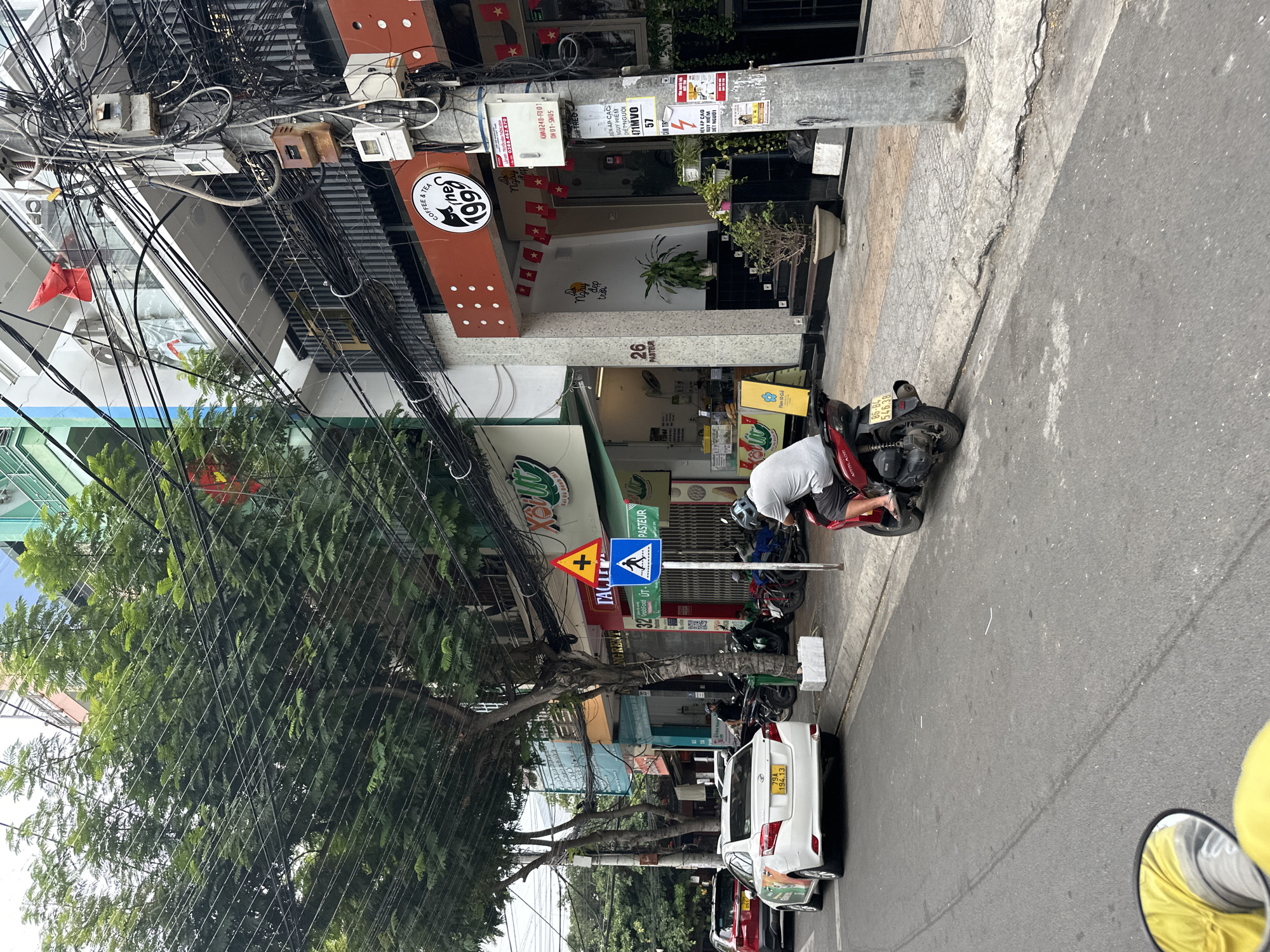 The image shows a street scene in Nha Trang Ward, Vietnam. Overhead, a tangle of power lines dominates the view. A man is tending to a parked red scooter on the sidewalk next to the street. Several parked scooters are further down the sidewalk, near a white car with license plate "79A 194.13". Street signs are present, including one indicating a pedestrian crossing. Buildings flank the street, with visible storefronts for coffee shops, food vendors ("Food ÚT"), and other businesses. A sign reads "26 PASTEUR". The street appears partially shaded by a large tree.