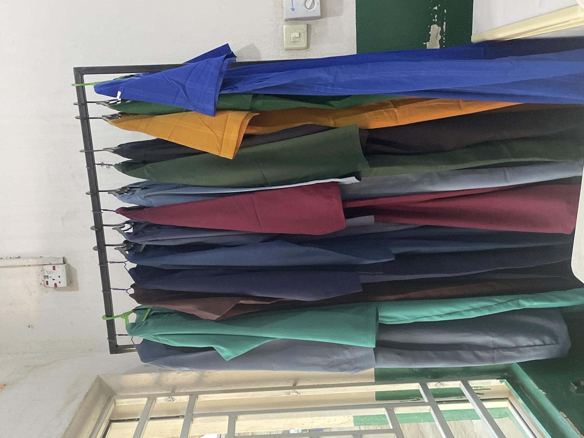 A rack of clothing in various colors hangs from a metal frame against a white wall. The items are folded and appear to be fabric or garments. The colors visible include royal blue, dark green, yellow, black, burgundy, navy blue, and a teal green. The scene is indoors, with a window and part of a green-painted wall visible in the background. There is an electrical outlet and a light switch on the white wall. The lighting suggests daytime. The overall impression is one of storage or inventory of fabric or clothing items.