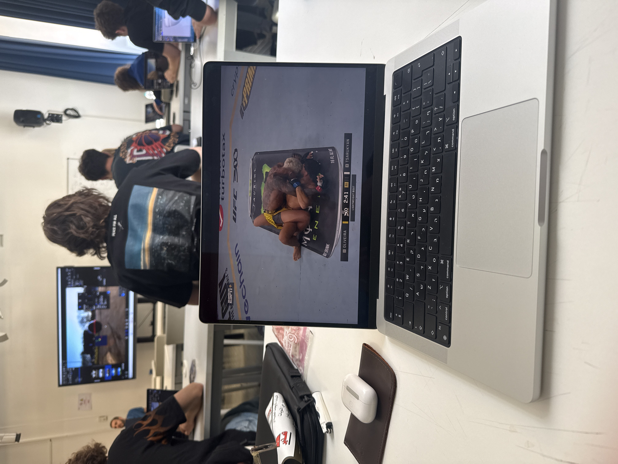 The image presents a classroom setting with several students. In the foreground, a silver MacBook Pro displays a UFC 300 fight on its screen. Adjacent to the laptop, there's a brown leather mat with a pair of white AirPods on it. A black laptop bag and a white object, possibly a shoe, are also visible nearby. Behind the laptop, a student with curly hair is wearing a black shirt with a graphic design. Further back, a large screen displays a video game. Other students are seated at tables, working on their laptops. The environment appears to be a modern classroom with white walls and a blue curtain. The location is indeterminable from the image.