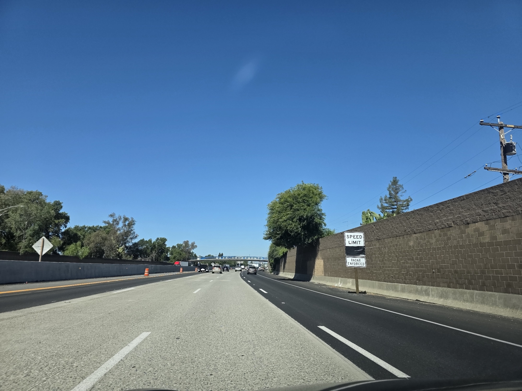 This image captures a perspective from inside a vehicle traveling on a highway in West Sacramento, California. The scene is set under a clear, bright blue sky, indicating a sunny day with good weather. The highway has multiple lanes, with other vehicles visible in the distance, suggesting moderate traffic.
To the right of the highway, a tall, textured brown brick wall lines the road, partially obscured by a large, leafy green tree. A signpost stands near the wall, displaying two white rectangular signs. The upper sign reads "SPEED LIMIT" and the lower sign states "RADAR ENFORCED."
On the left side of the highway, there is a concrete barrier with orange traffic cones visible in the distance, suggesting possible construction or maintenance. Beyond the barrier, a line of trees and foliage separates the highway from what appears to be a residential or commercial area. A white diamond-shaped sign with a reflective surface is also visible on the left.
In the far distance, an overpass with text that appears to read "WEST SACRAMENTO" can be seen, confirming the location. Overhead power lines and a utility pole are visible against the sky on the right side of the image. The overall impression is one of routine travel on a sunny day in a suburban highway environment.
