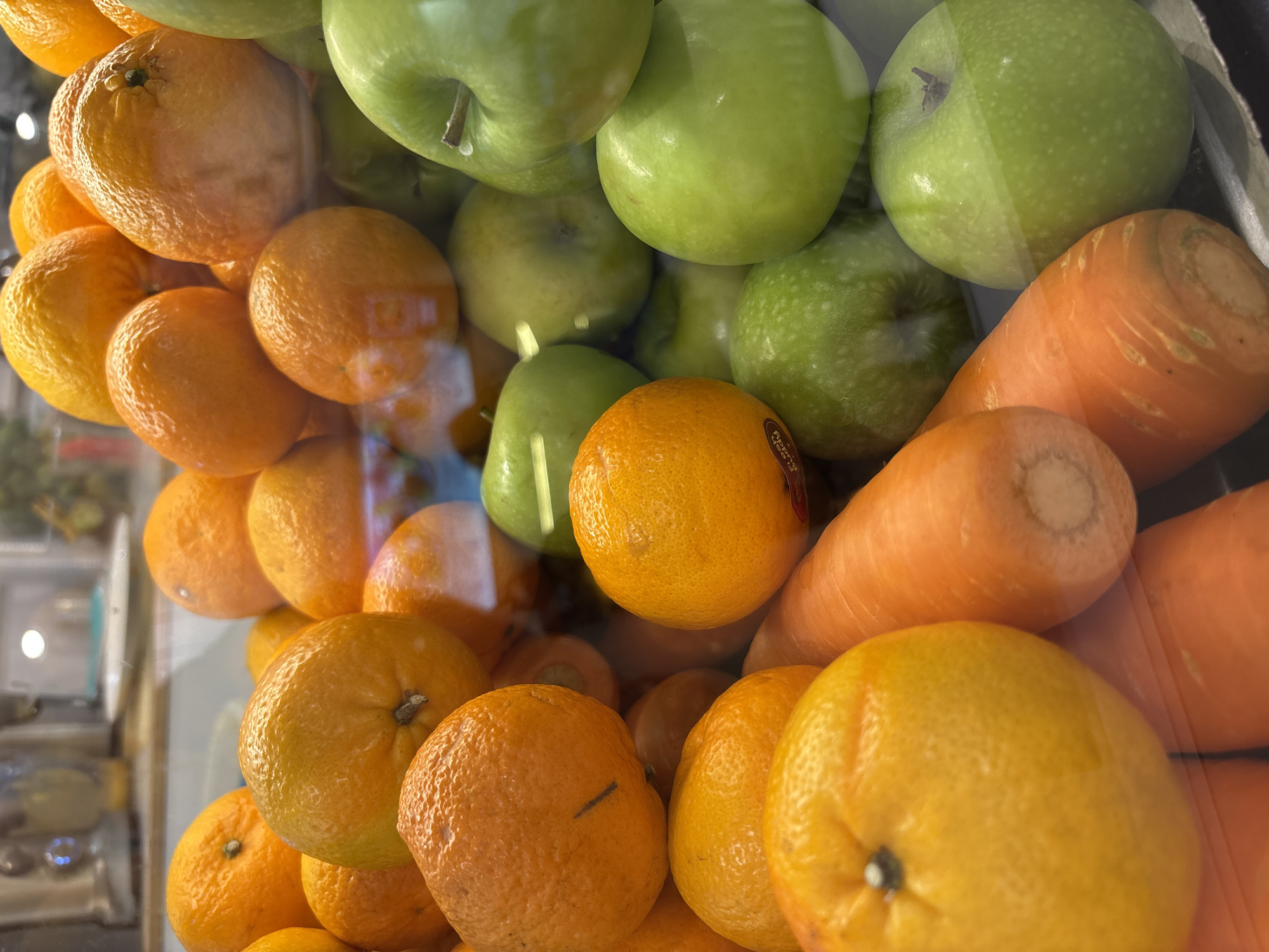 The image is an eye-level close-up shot of fresh produce behind glass, possibly a display case, located in an unknown location in the city of บ้านบางรักษ์, Thailand. The composition includes a pile of oranges with visible peels, a cluster of green apples with some stems visible, and two orange carrots. The produce appears to be packed together tightly. Reflections are visible on the glass, hinting at the surrounding environment.