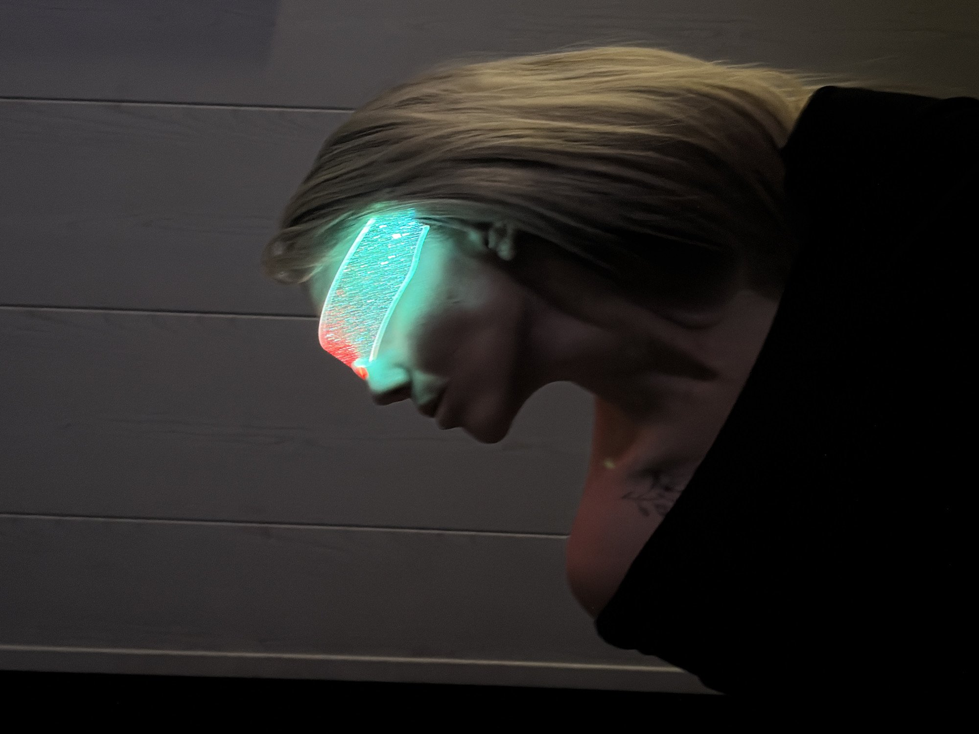 A person with blonde hair is depicted in profile view. The individual is wearing a black garment. A glowing, rectangular object is positioned over their forehead, emitting green and red light. A tattoo is visible on their upper chest or shoulder. The background consists of a wall with horizontal planks, likely wood. The location is not discernible from the image.