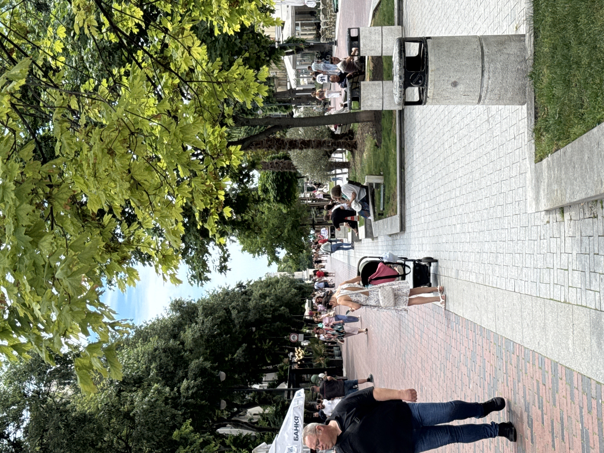 The image depicts a lively pedestrian street in Varna, Bulgaria, likely along the Sea Garden promenade, judging by the presence of mature trees and the general atmosphere. The foreground features a man in a black polo shirt and blue jeans walking on a red brick pathway. To his right, a woman in a white floral dress pushes a stroller on a grey paved sidewalk. In the background, a diverse group of people are strolling, sitting on benches, and patronizing outdoor cafes. Lush green trees line both sides of the street, creating a canopy effect. Several outdoor seating areas with tables and chairs are visible, suggesting a vibrant commercial area. The overall scene is one of a bustling urban environment on a pleasant day, with ample green spaces and amenities for pedestrians.