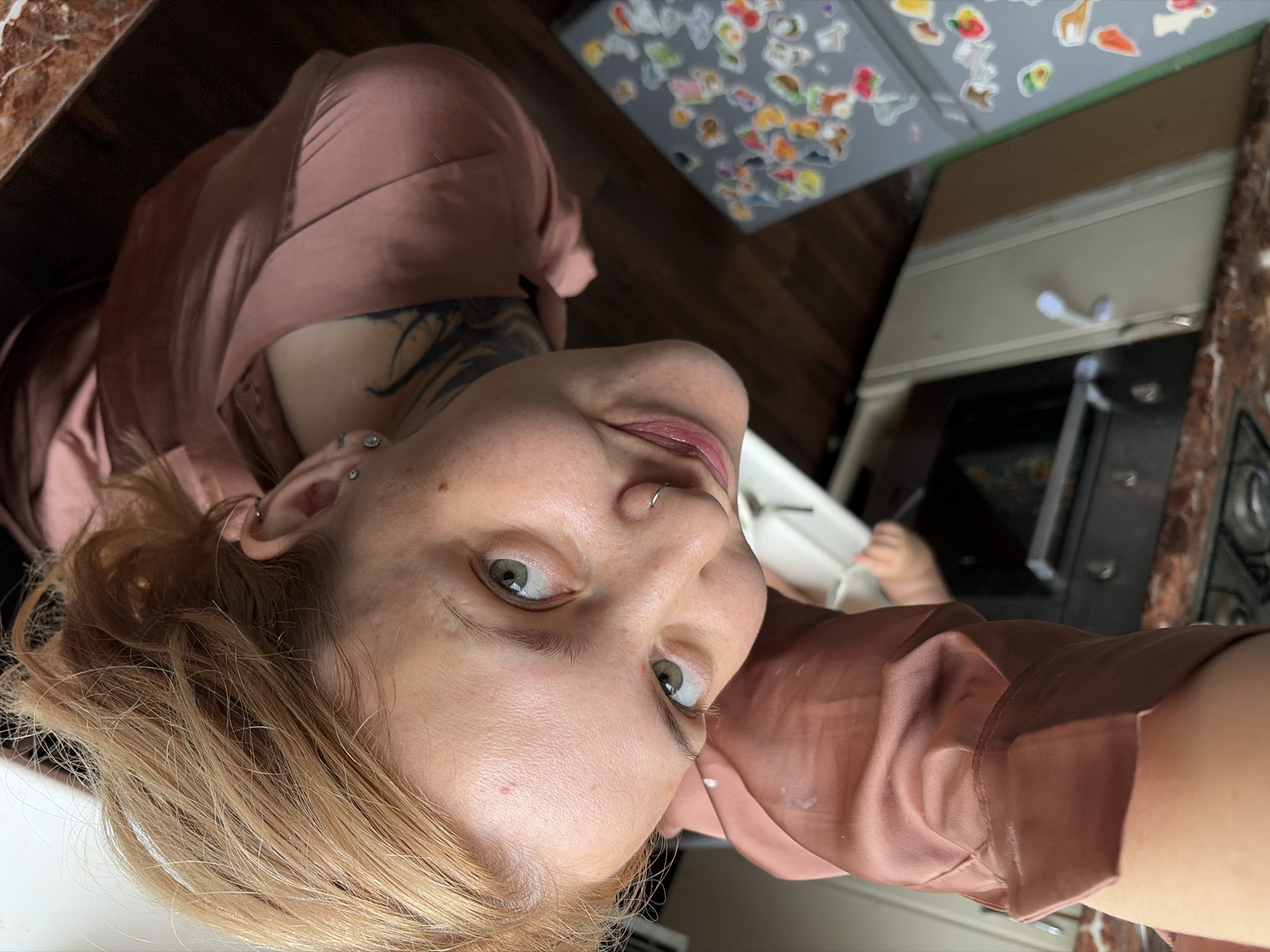 A woman with blonde hair is positioned in a kitchen, wearing a reddish-brown satin top. She has piercings on her ear and nose, with tattoos visible on her upper chest and neck. The kitchen includes an oven, countertop, and refrigerator covered in stickers. The location is within the city of Chaikovsky, Russia, which is inferred from the presence of common household items and kitchen appliances typically found in residential settings. The subject is facing the camera.