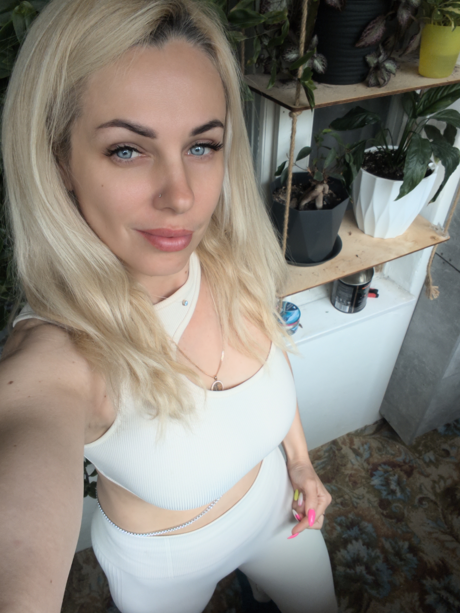 A blonde woman with blue eyes is taking a selfie in what appears to be an indoor setting, potentially in an apartment or house. She is wearing a white, two-piece outfit consisting of a crop top with a cut-out design and matching leggings. She has a silver chain around her waist, a necklace, and a nose piercing. Her nails are painted in various bright colors.
Behind her, there's a shelf unit with various potted plants. A white cabinet is also visible. The floor has a patterned rug.
The location in the city of Sevastopol, Ukraine, cannot be determined from the image.