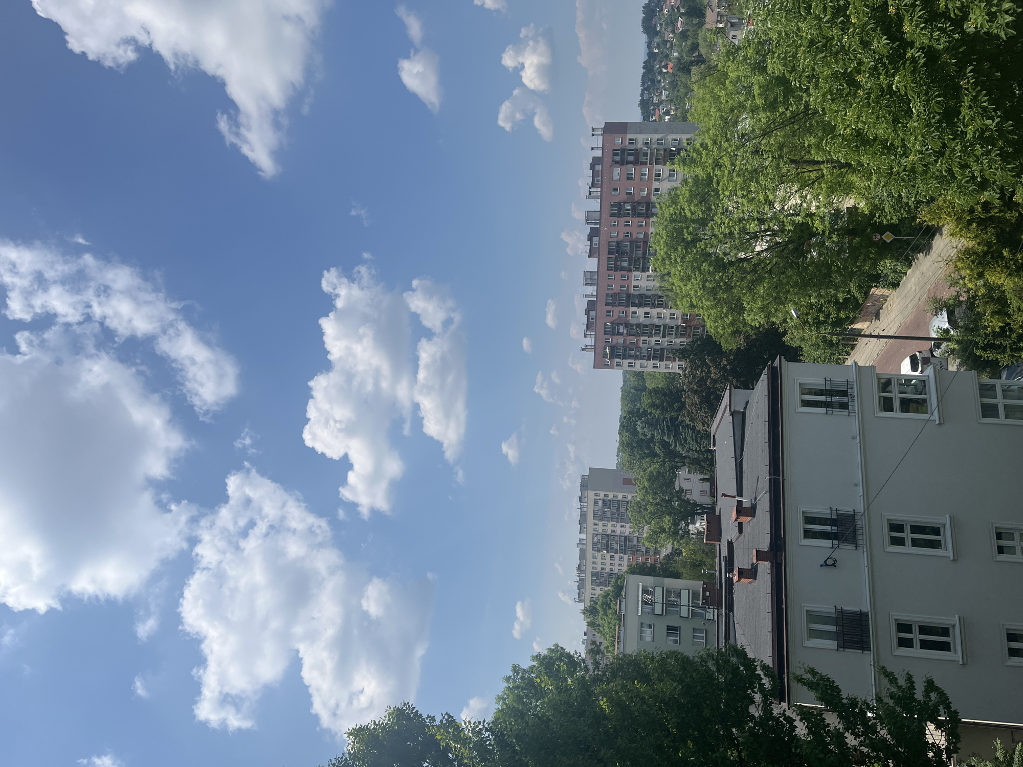 The image presents a vibrant urban landscape on a clear, sunny day, captured from an elevated viewpoint, possibly a window or balcony. The upper portion of the frame is dominated by a bright blue sky, adorned with scattered, fluffy white cumulus clouds.
In the mid-ground, a prominent multi-story apartment building with a reddish-brown and lighter-toned facade stands tall, featuring numerous windows and balconies. Further in the distance, a residential hillside densely covered with houses and greenery can be seen.
The immediate foreground includes the rooftops and upper levels of a white building, displaying several windows, some of which are equipped with protective metal grilles. Abundant lush green trees frame the right side and bottom of the scene, and are also interspersed throughout the urban setting. Partially visible through the foliage on the right is a paved street or road, where a white car is parked. A street lamp and a yellow diamond-shaped road sign, characteristic of a European "priority road" or similar regulation, are also discernable.
The scene depicts a calm daytime setting under fair weather conditions. There are no visible people or specific activities taking place. The architectural style of the buildings and the design of the road sign suggest a European city, potentially in Central or Eastern Europe. No explicit text is readable within the image.