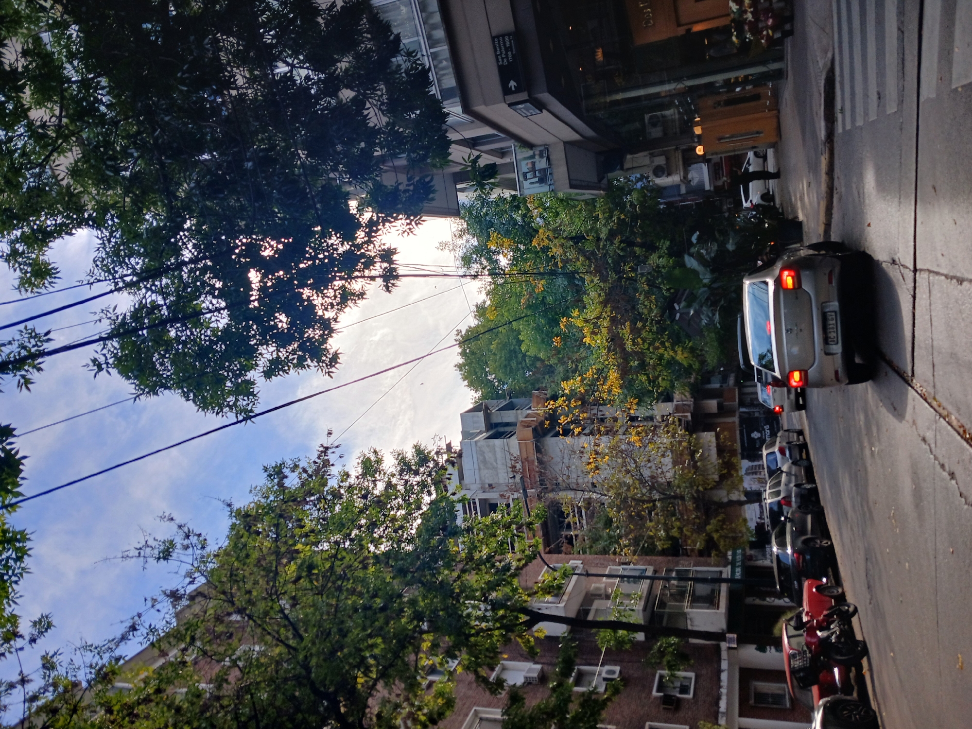A street scene in Buenos Aires, Argentina, viewed from a low angle. Buildings line both sides of the street, partially obscured by trees. Power lines are visible against a blue sky with scattered clouds. Cars and motorcycles are parked along the right side of the street. There is a pedestrian crossing, and people can be seen in the background near a building. A street sign reading "San Benito de Palermo 1700-1800" is visible. The overall scene suggests an urban residential area.