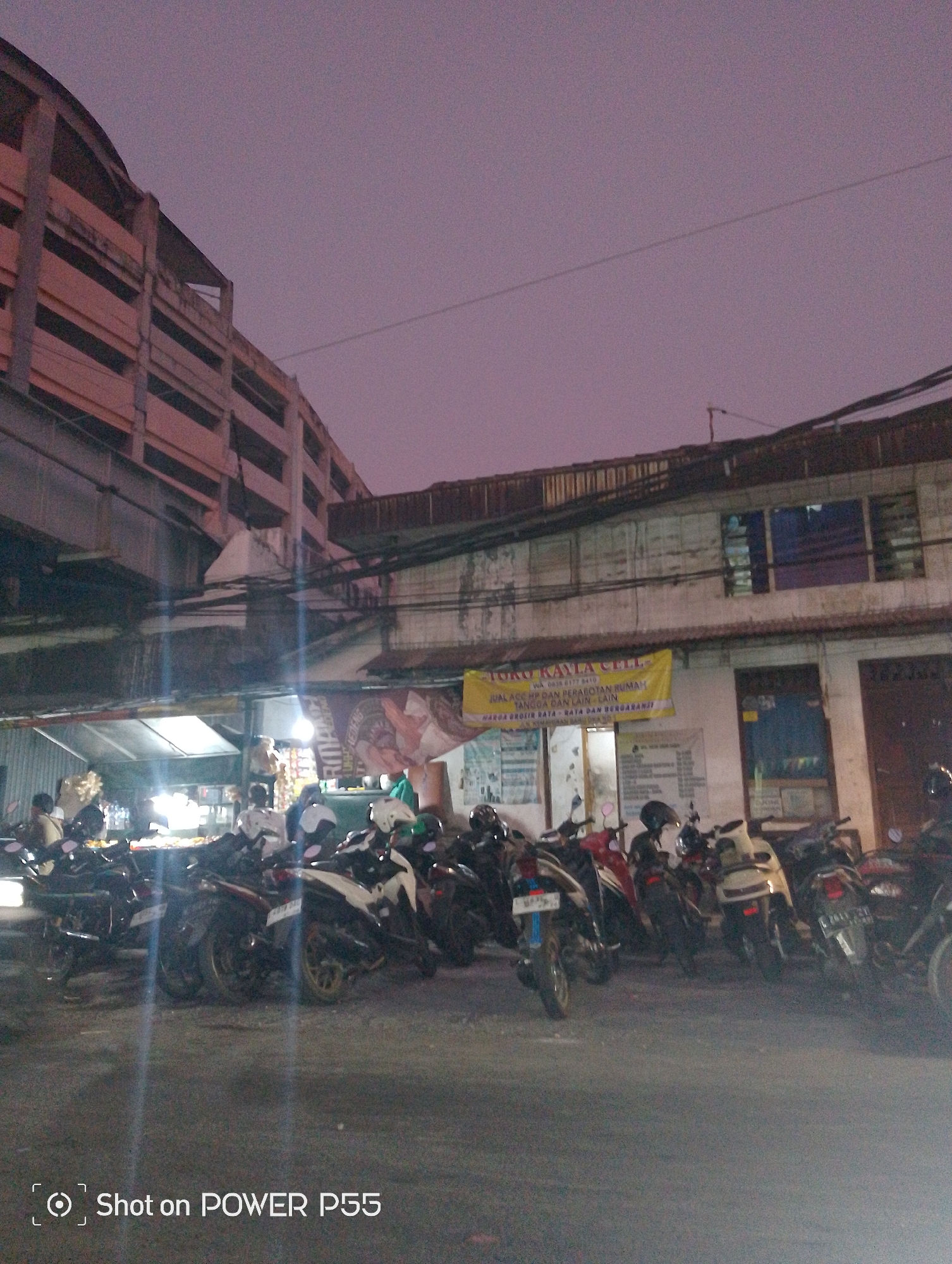 This image depicts a street scene at dusk or dawn, likely in Surabaya, Indonesia, given the presence of motorcycles and the overall urban setting. In the foreground, a row of parked motorcycles is visible. Behind the motorcycles, a low-rise building with a tin roof is present. A banner hangs from the building, advertising "TOKO KAYLA CELL" and listing phone numbers and product offerings, including mobile phones and home appliances. The building appears to be a shop or a small business. Further in the background, a large, multi-story concrete structure, possibly a parking garage or an elevated building, looms over the scene. The sky is a muted purple and pink, indicating low light conditions. A few individuals are standing near the shop, possibly customers or employees. The environment suggests a commercial area within the city.