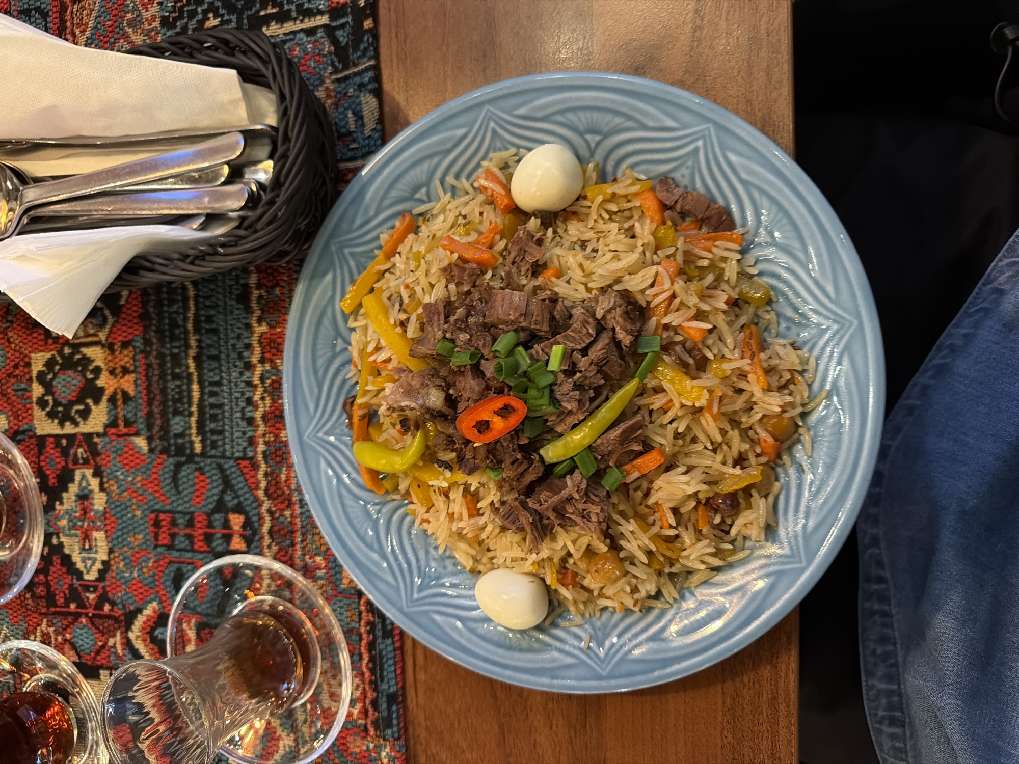 The image depicts a plate of plov, a rice dish, on a wooden table. The plov consists of rice, meat, carrots, yellow peppers, raisins, and is garnished with green onions and a red chili pepper. Two peeled quail eggs are placed on the plate. The plate is light blue with a decorative pattern. Beside the plate is a basket filled with silverware and napkins. Next to the basket are three armudu glasses, two of which are filled with tea. A patterned rug lies under the basket and glasses. The edge of a pair of denim jeans is visible on the right side of the frame. The setting appears to be a restaurant in Warsaw, Poland based on the common presence of plov in that city.