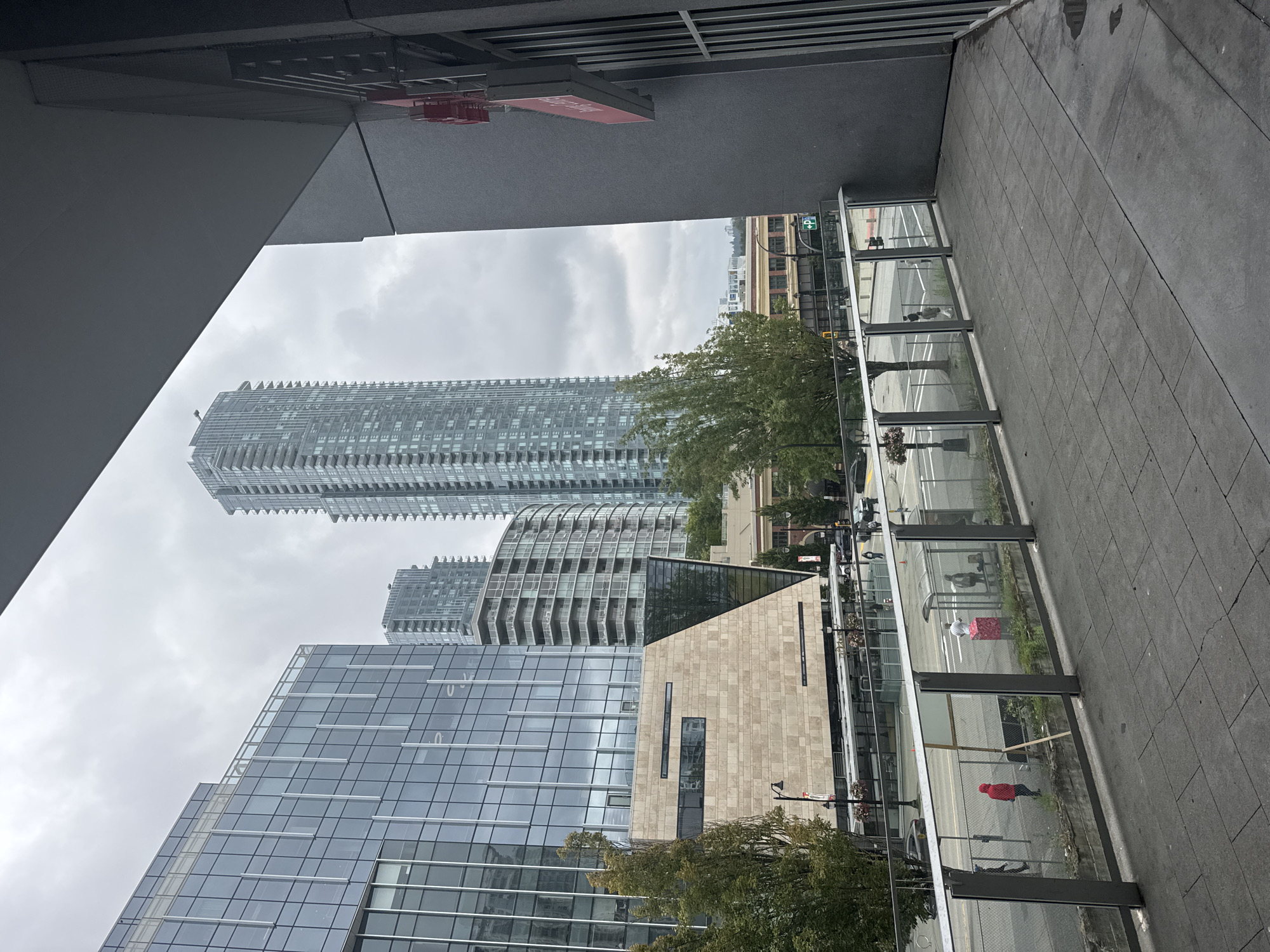 This is an outdoor, eye-level, medium shot photograph, captured on an overcast day. The scene depicts a modern urban environment in New Westminster, Canada, with a focus on contemporary architecture.
In the foreground, a paved concrete walkway with square-patterned slabs leads to a glass-railed balcony. Beyond the railing, a street is visible with cars and pedestrians. A bus shelter with a transparent roof and side panels is situated on the sidewalk, and several individuals are standing or walking near it. One person in a red jacket stands out against the muted background.
The mid-ground features a street with visible lane markings and traffic signals. Trees with green foliage line the street, and streetlights are also present. A light-colored building with a textured stone facade dominates the mid-left portion of the image, with a sharp, geometric glass section integrated into its design. Behind this building, taller modern structures rise, including a distinctive, slender, light-colored skyscraper with numerous windows and balconies. Another, wider, light-colored building with rounded balconies is visible further back.
The background consists of a cloudy, gray sky and more distant urban buildings. On the right side of the image, a dark gray building with vertical window-like elements and a prominent red sign is partially visible, framing the scene. The overall atmosphere is one of a developed city district with a mix of office and residential buildings.