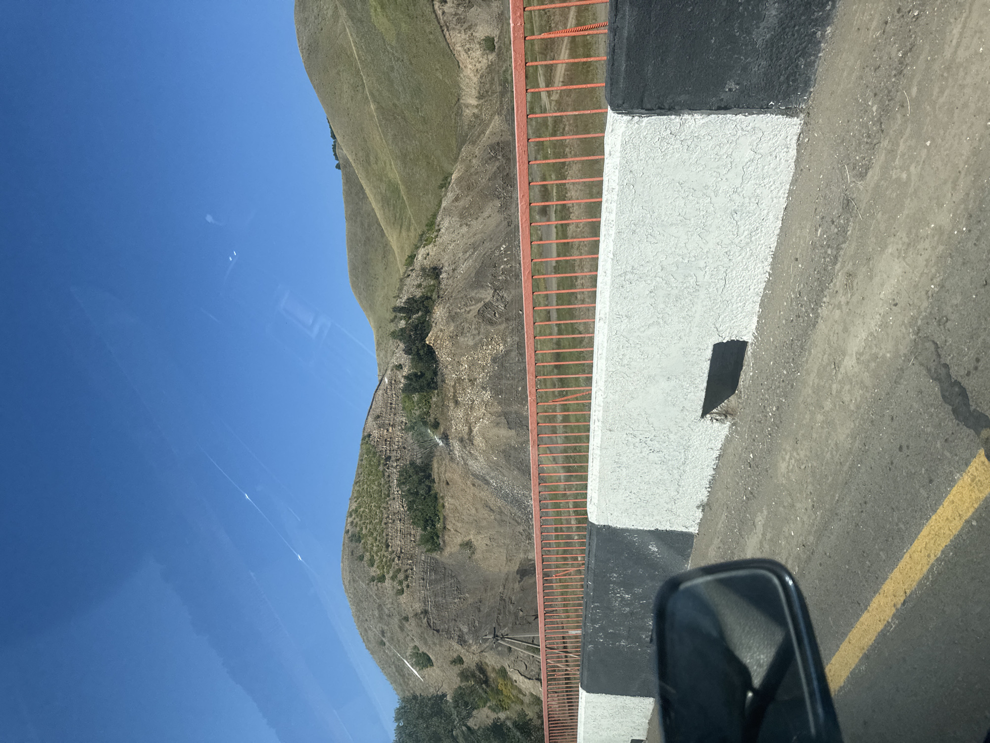 The image depicts a view from a moving vehicle, likely on a bridge, looking towards a hilly, barren landscape under a clear blue sky. In the foreground, a roadside barrier is visible, painted with alternating white and black sections, and a bright orange railing. A yellow lane marker is present on the asphalt road. To the left, a side mirror of the vehicle is partially visible. The background features rolling hills with patches of green vegetation and exposed rocky areas, suggesting a rural or mountainous environment. There are no people or animals in the frame, and the scene appears to be a still moment captured during transit, possibly on the road leading into or out of Kugarchi, Russia, given the implied location.