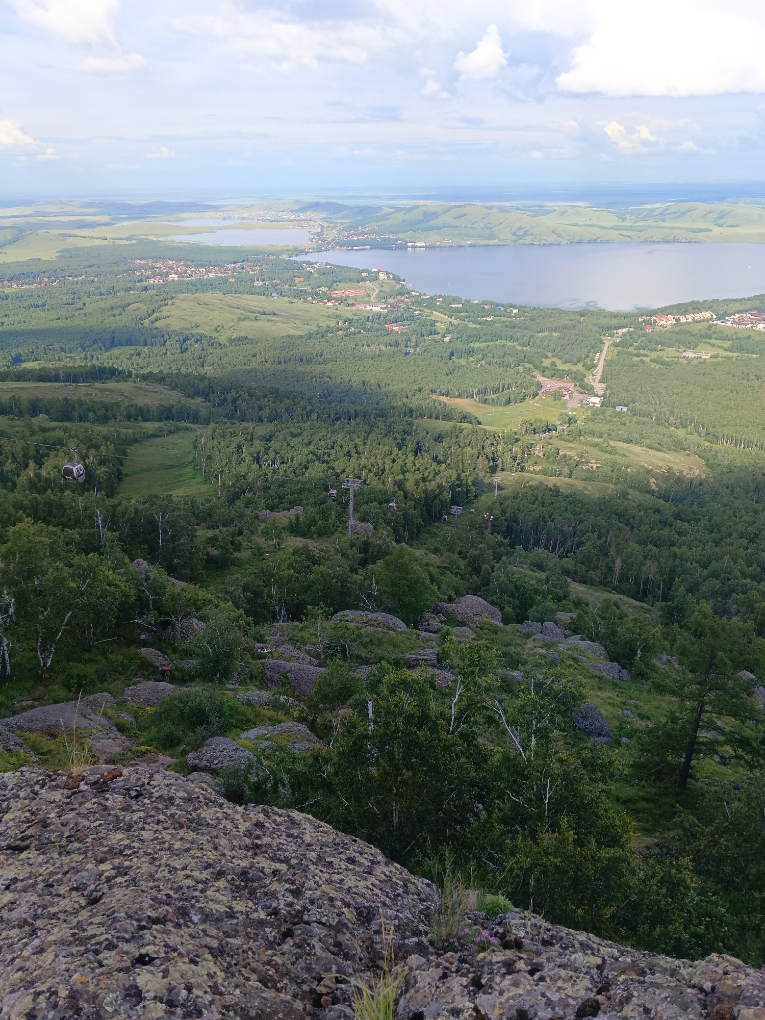 An aerial view captures a sprawling landscape with a village nestled between a forest and a large body of water. The foreground is dominated by a rocky, moss-covered slope. In the middle ground, a dense forest stretches across rolling hills, punctuated by a ski lift system with visible cabins and towers. Beyond the forest, a settlement with numerous buildings is spread out along the shoreline of a calm lake. The background features a hazy horizon where the lake meets distant, undulating hills under a sky with scattered clouds.
