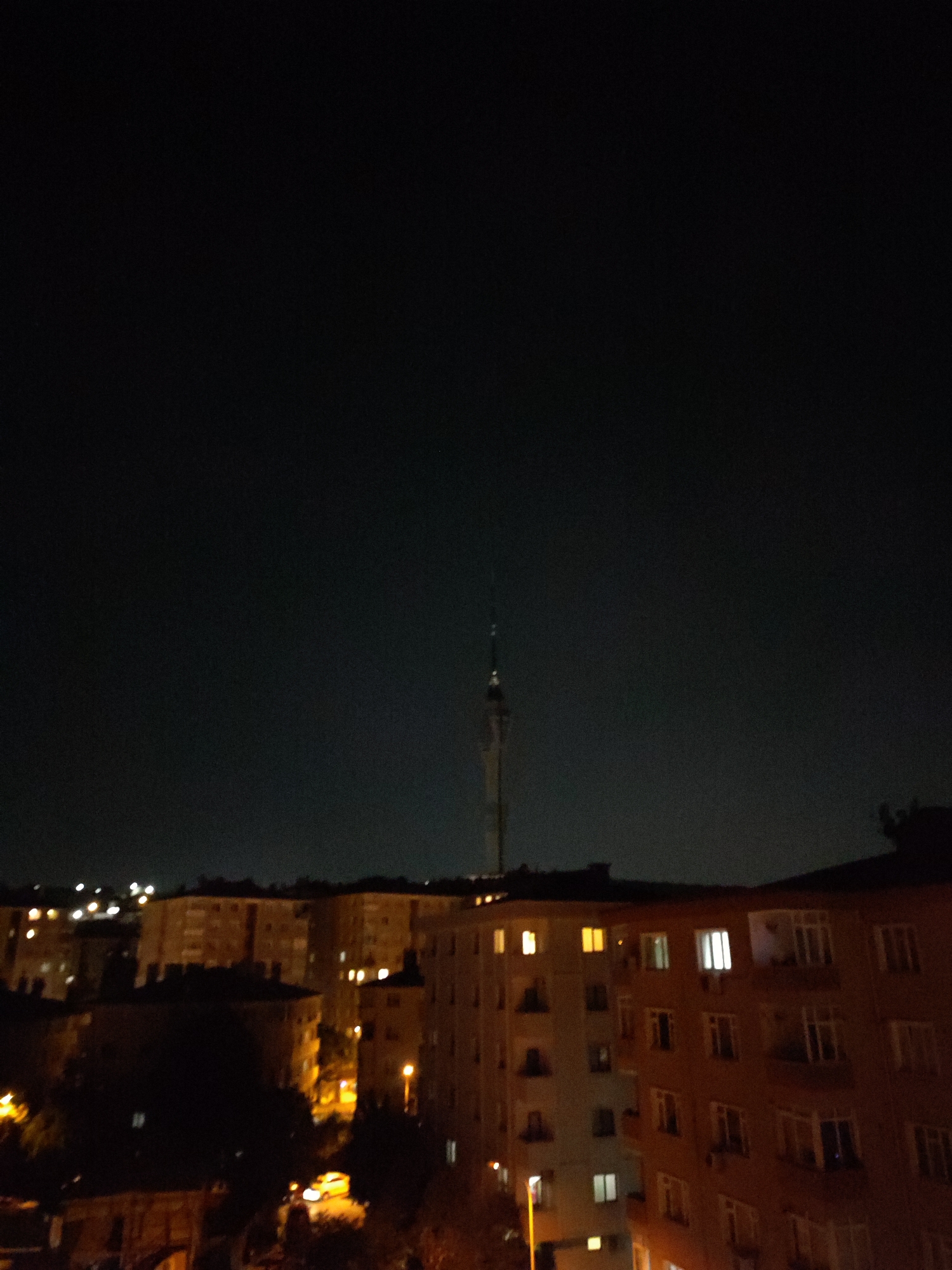 The image is a nighttime aerial view of a city skyline in Üsküdar, Turkey. The sky is a deep black, with a tall, slender tower with a light at its peak visible in the upper center of the frame. Below the tower, the lights of apartment buildings create a warm glow against the dark night. The buildings are multi-story structures with visible windows, some illuminated from within. In the foreground, darker silhouettes of trees and other structures are present, hinting at dense urban development. A streetlamp illuminates a patch of ground in the lower right portion of the image. The overall impression is one of a quiet, illuminated urban landscape at night.