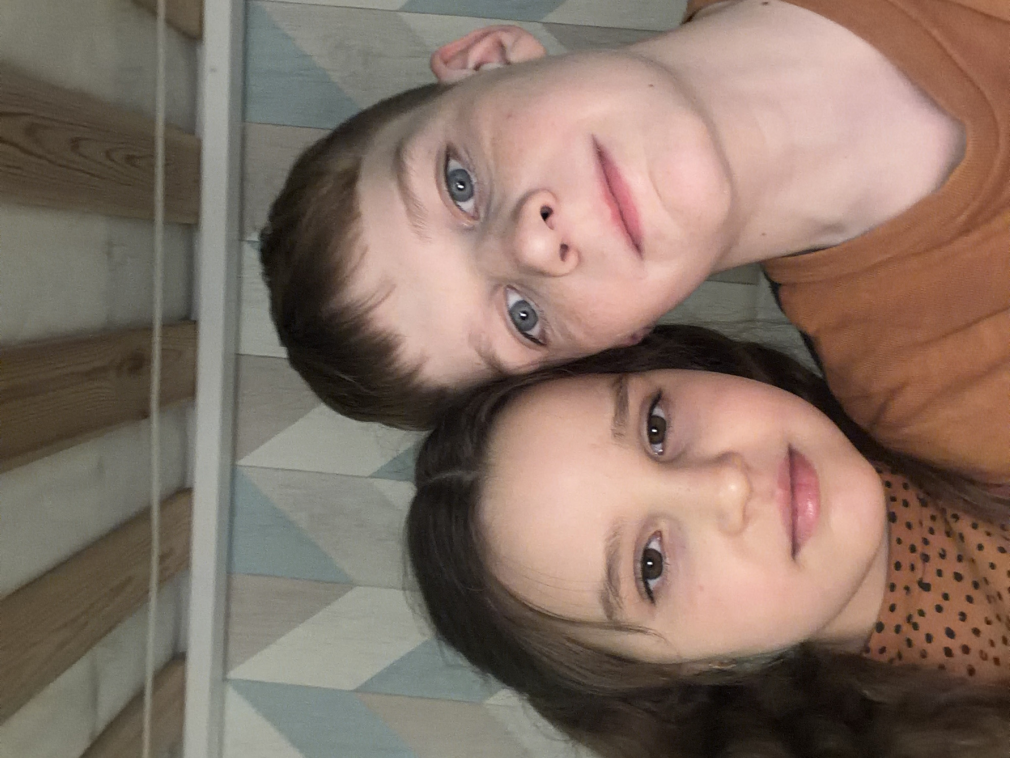 Two children are lying side-by-side looking at the camera. One child appears to be a boy with blue eyes, short brown hair, and is wearing an orange shirt. The other child appears to be a girl with brown hair, brown eyes, and is wearing a brown patterned shirt. The background appears to be a wall with a light geometric pattern. The location is unknown and there is no information to suggest it is in Zaprudnya, Russia.