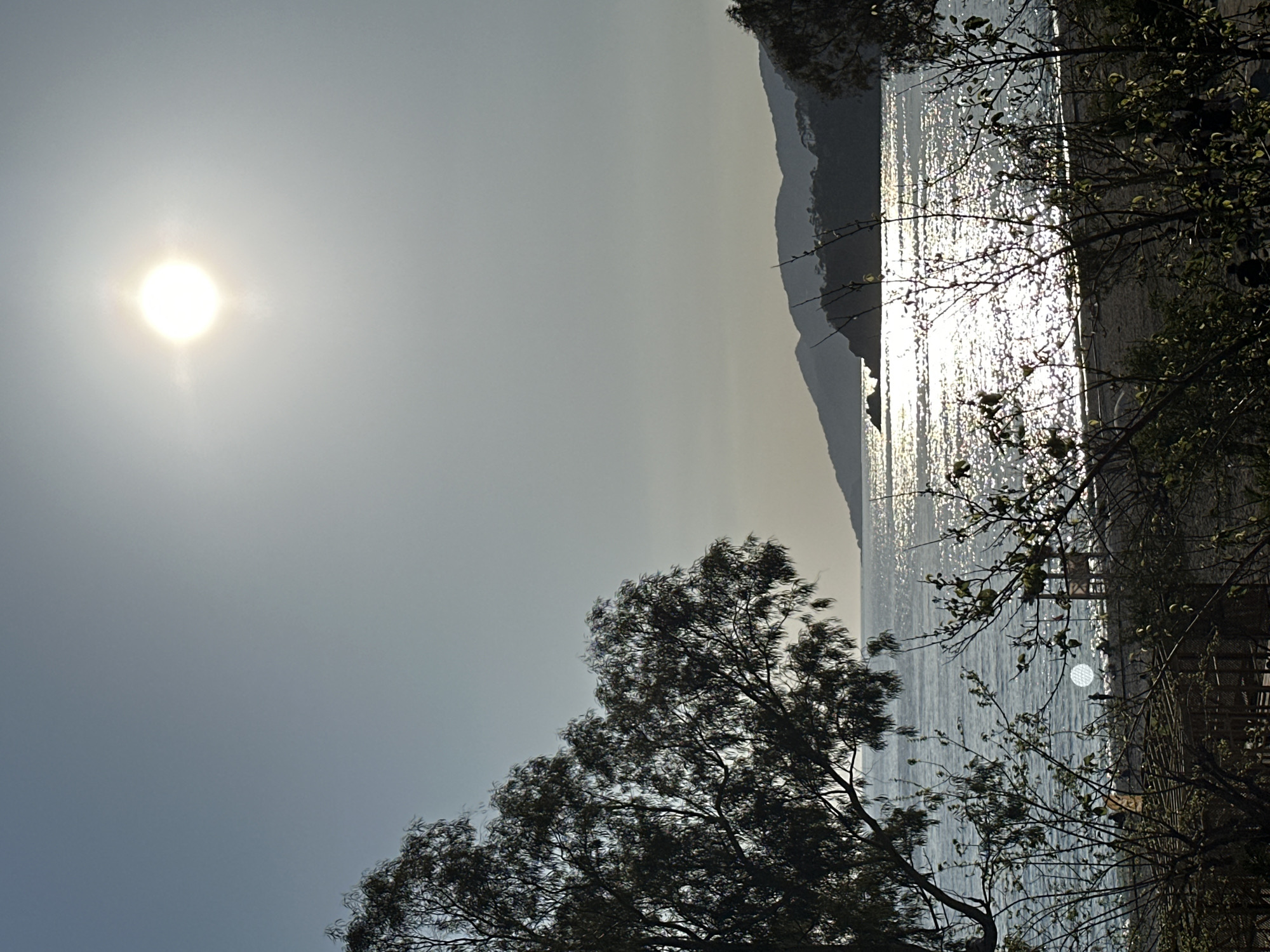 A hazy, bright sun shines over a body of water reflecting sunlight, framed by silhouetted mountains in the distance and dark trees in the foreground. The location is likely in Fethiye, Turkey, known for its coastal views and mountainous terrain. The image suggests a time of day with intense sunlight, possibly early morning or late afternoon. There are no people visible in the image.