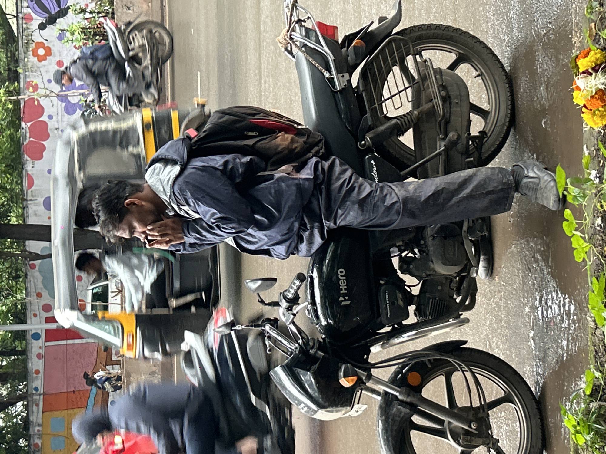 In Pune, India, on a wet, overcast day, a man in a dark blue hooded jacket and matching pants sits on a black Hero motorcycle. He is looking to his left and appears to be holding something to his mouth. The motorcycle has a silver gas tank with the "Hero" logo. The man wears grey shoes and a black backpack is strapped to his back. In the background, an auto-rickshaw, also in motion, is visible. The street is wet, with puddles reflecting the overcast sky. A colorful mural depicting flowers and a butterfly adorns a wall in the background. Green plants are visible in the foreground.