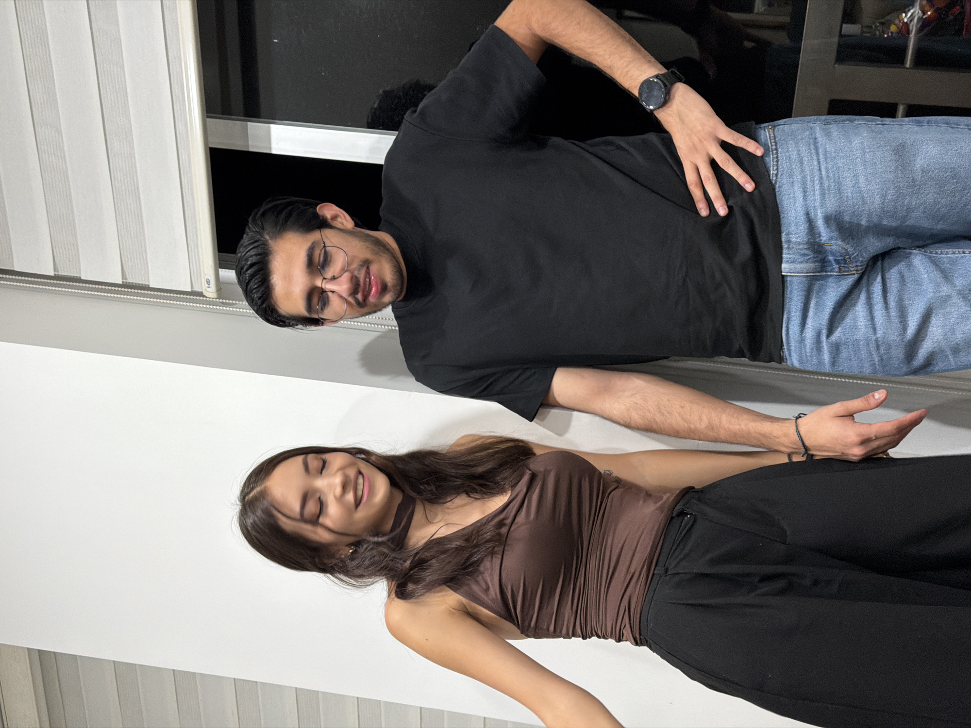 The image shows a man and a woman lying on a surface, possibly a floor, indoors. The man is wearing a black t-shirt, blue jeans, and a watch. He has glasses and dark hair. He is lying on his side and has his hand on his stomach. The woman is wearing a brown top and black pants. She has long brown hair. She is lying on her back with her eyes closed. The background includes a white wall and a window with blinds. The location is San Andrés Cholula, Mexico.