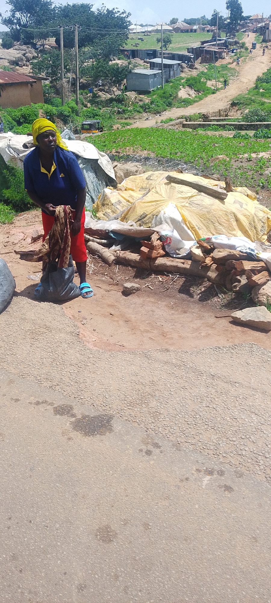 A woman wearing a yellow headscarf, a blue and yellow polo shirt, and red shorts stands on a dirt path. She is holding a patterned cloth, and a black plastic bag is at her feet. The background shows a dusty, rural landscape with scattered buildings, some made of corrugated metal and others of mud or stone. Vegetation is sparse in some areas, with green crops visible in the middle ground. A dirt road winds through the scene, with a few people and a yellow tricycle visible in the distance. The ground in the foreground is paved with asphalt that has oil stains. The scene appears to be a residential area in Jos, Nigeria.