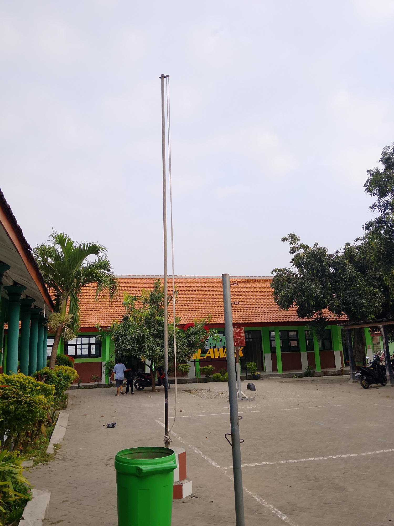 The image depicts a schoolyard in the city of Kepuh, Indonesia. In the foreground, a tall, empty flagpole stands to the right of a bright green trash can. The ground is paved with interlocking gray bricks. To the left, a building with a red-tiled roof and green trim runs along the edge of the frame, featuring a row of large, dark green columns. Beyond this building, a palm tree and other green foliage are visible. In the middle ground, another building with a red-tiled roof and green walls is visible, with several windows and some signage that reads "SPU LAWAK" in yellow letters. Two people, one wearing a blue shirt and shorts, and another in dark clothing, are walking away from the camera toward the building. A motorcycle is parked near the building. The sky is overcast and a pale gray.