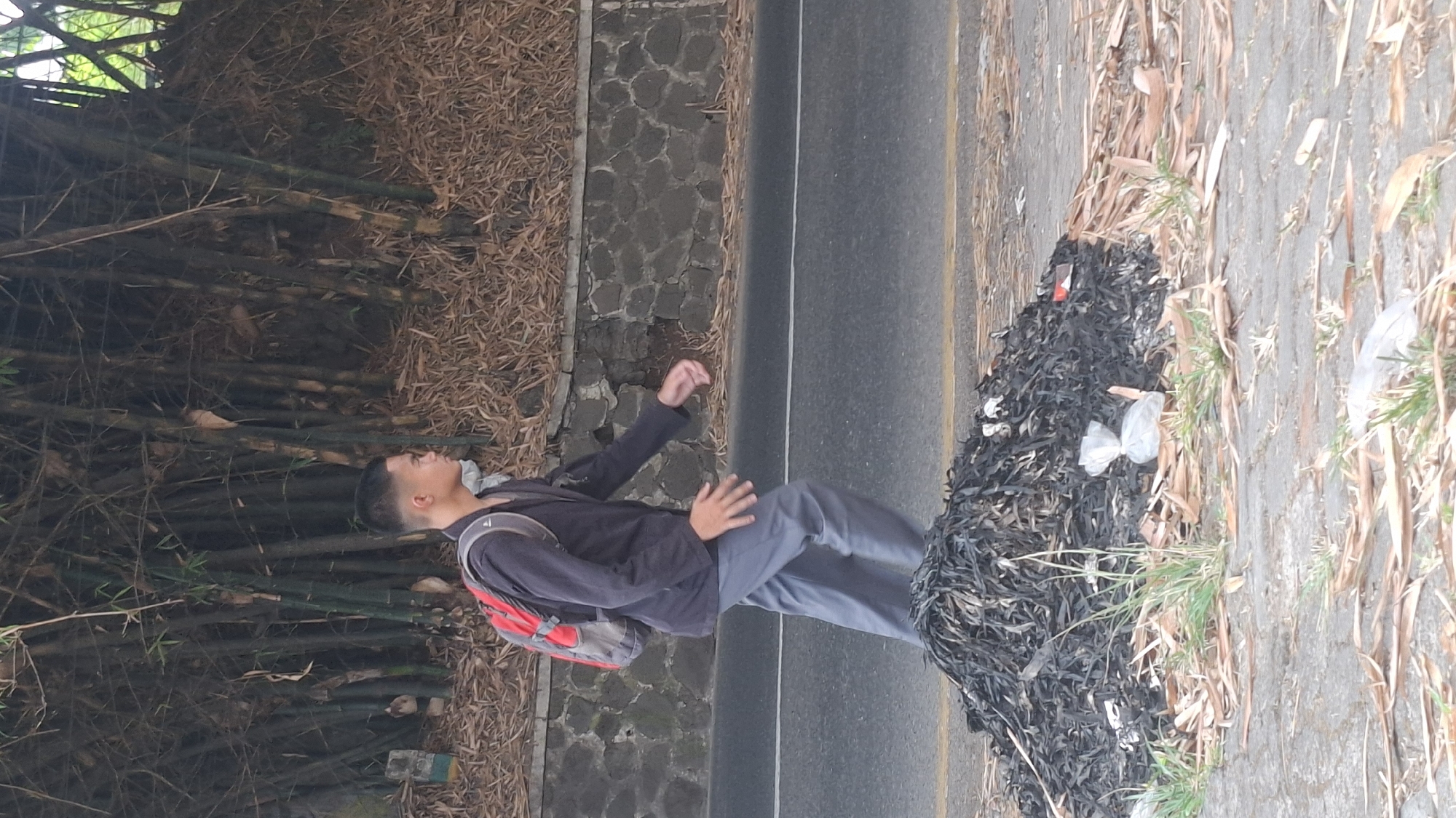 A young man with short hair, wearing a dark jacket and grey pants, walks along a paved road bordered by a stone wall and bamboo trees in Cilaku, Indonesia. He carries a red and grey backpack. In the foreground, a pile of dark, shredded material, possibly dried leaves or rubber, is present on the roadside. Scattered dry leaves and patches of grass are visible on the paved area next to the road.