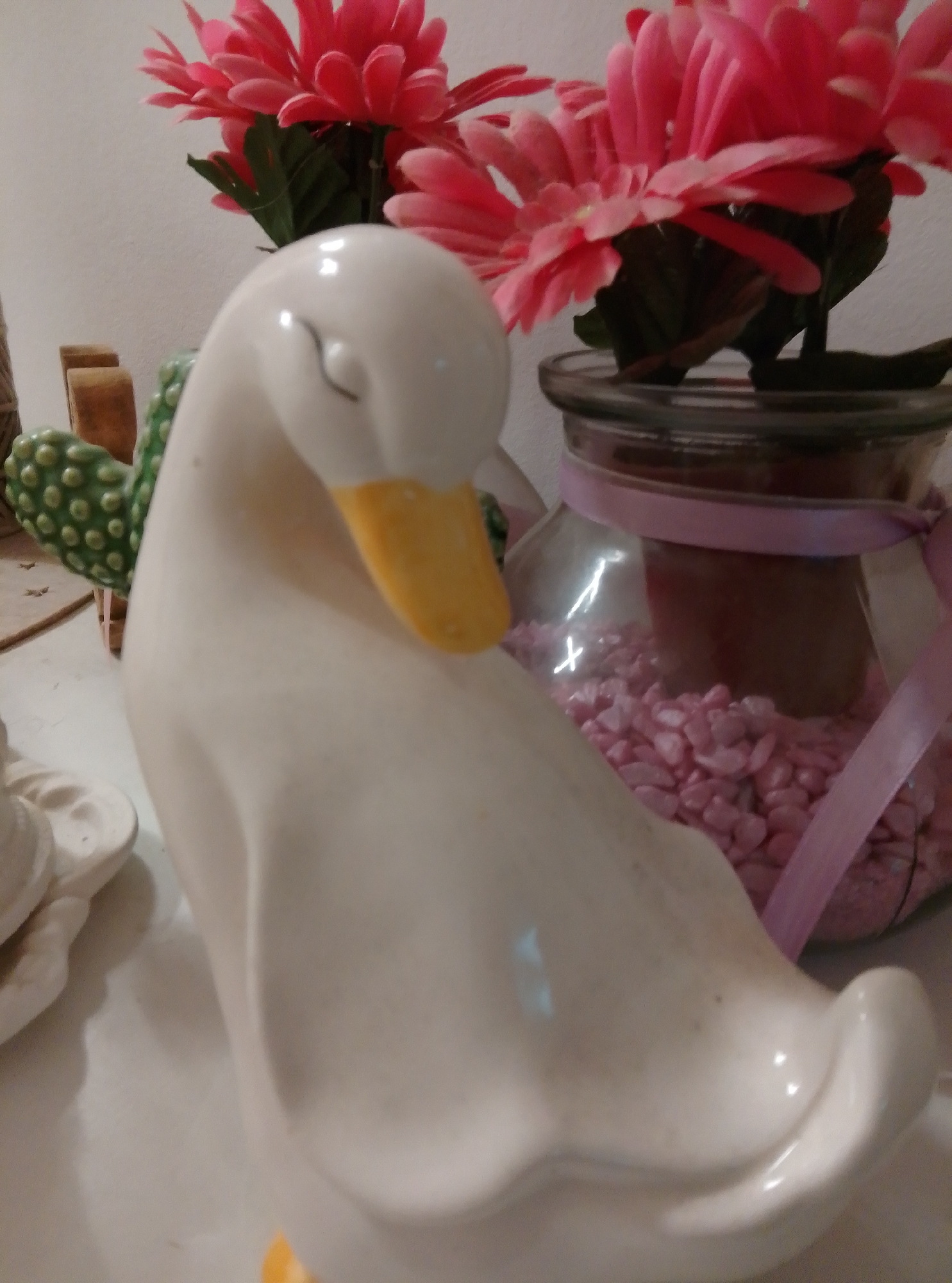An ornamental white ceramic duck with a yellow beak is positioned in the foreground, facing to the right and slightly downward. Behind it, a glass jar filled with small pink stones and a dark object is visible, tied with a pink ribbon. To the left of the duck, a green ceramic cactus decoration with raised bumps is partially visible. In the background, bright pink gerbera daisies are arranged in a bouquet. The scene appears to be indoors, with a plain white wall as the backdrop. There is no indication of the city of Wedel, Germany, or any specific location.